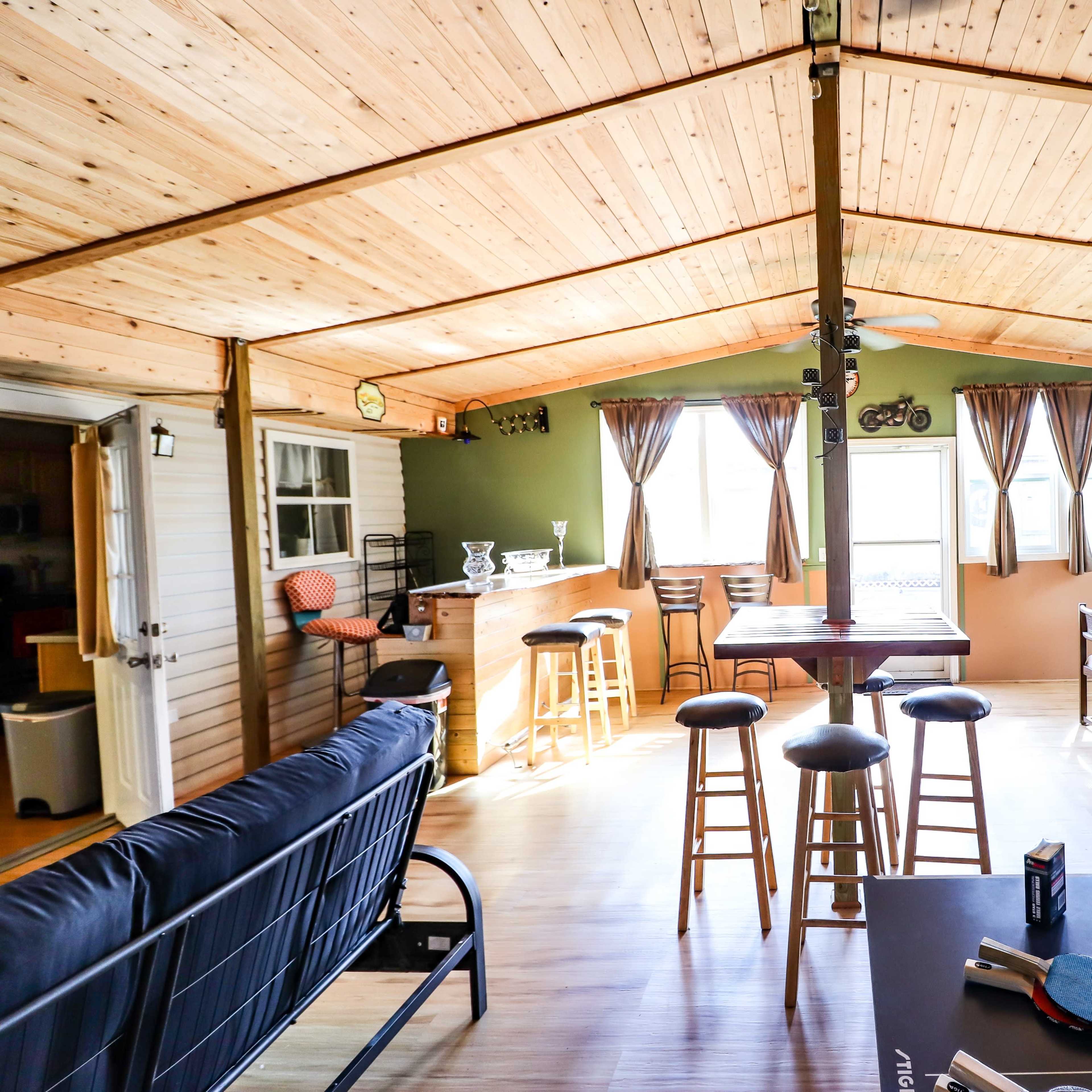 A bright, spacious room with wooden ceilings, a bar area, seating, and large windows letting in natural light.