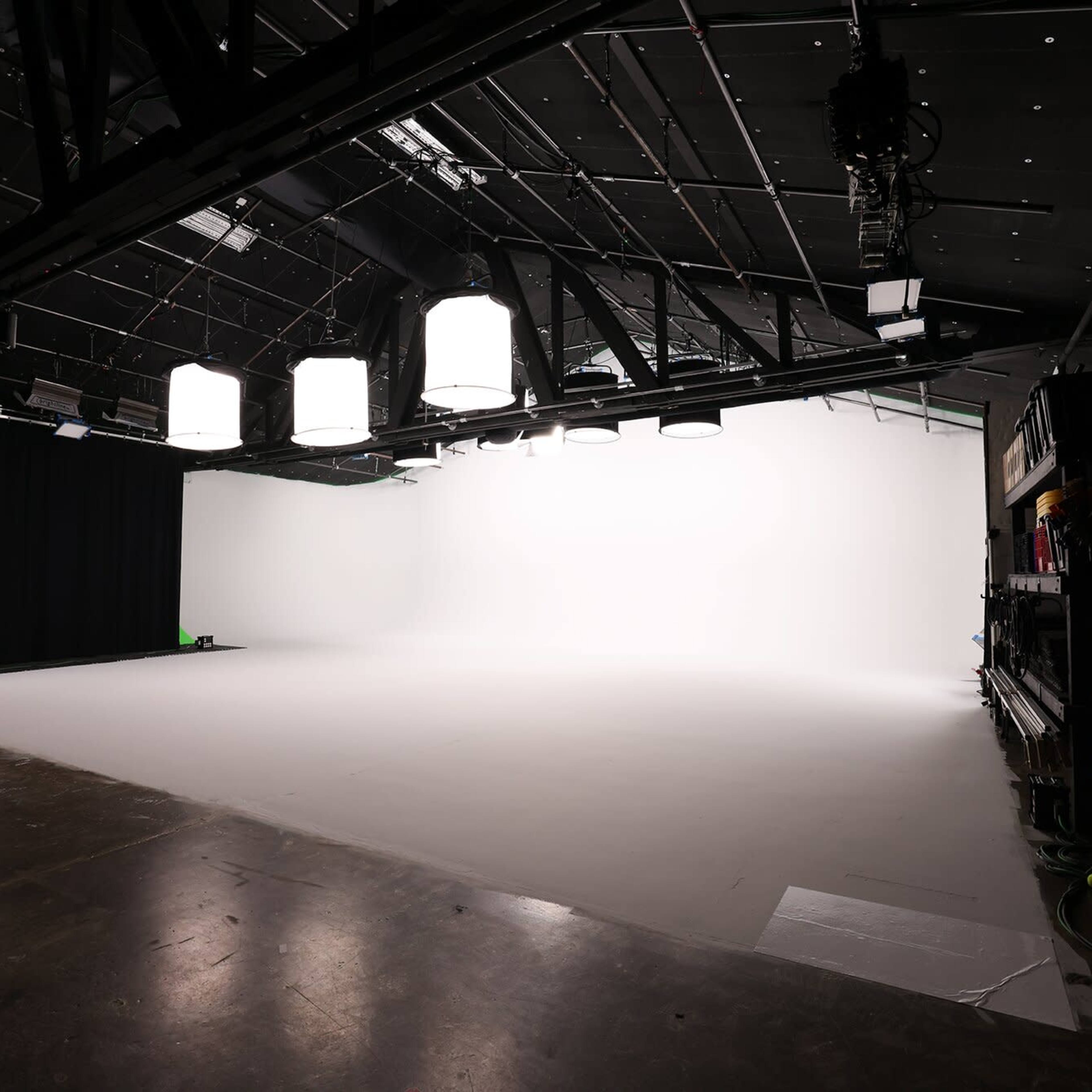 A spacious film studio with a white backdrop and professional lighting equipment on the ceiling.