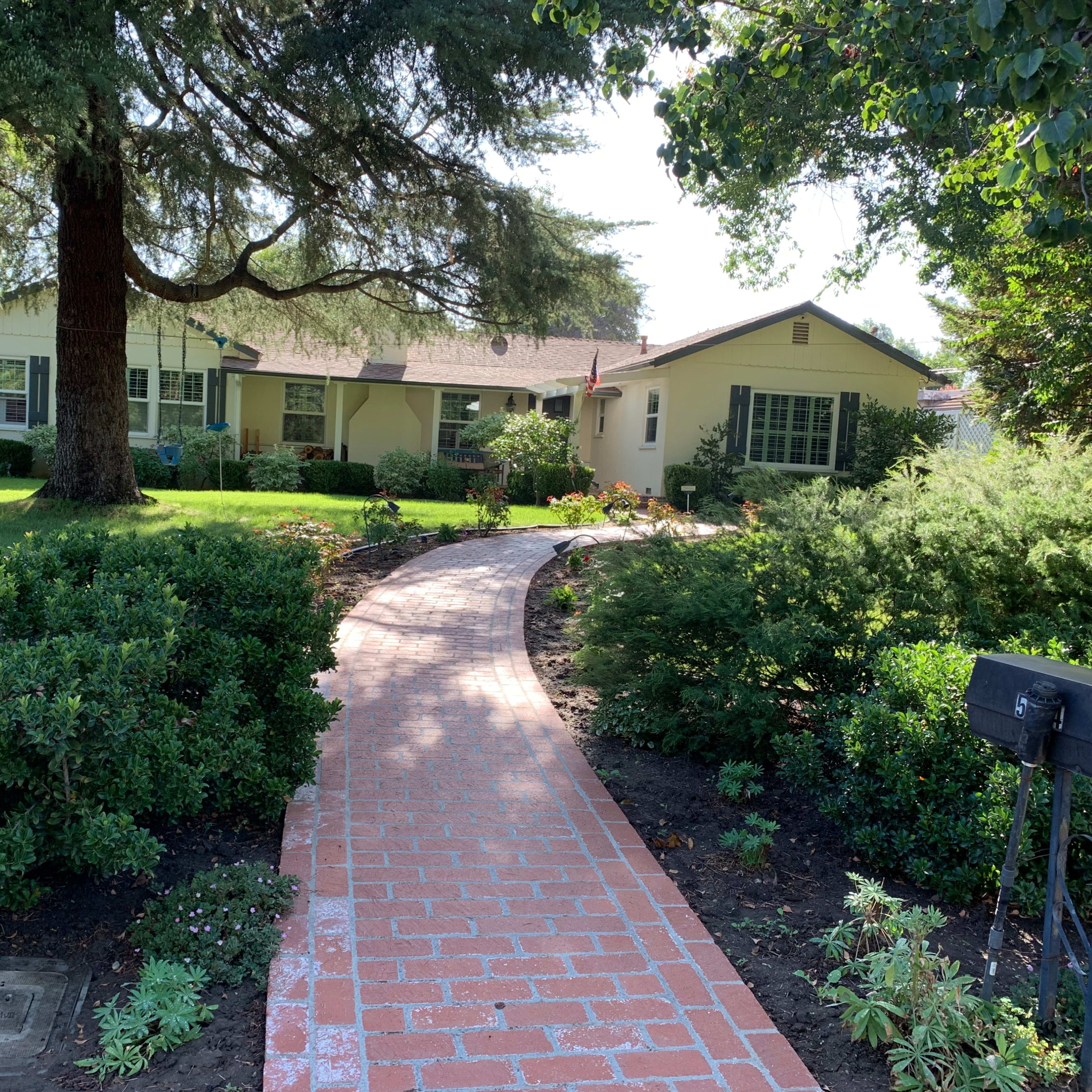 A brick pathway leads to a single-story house surrounded by well-maintained greenery.
