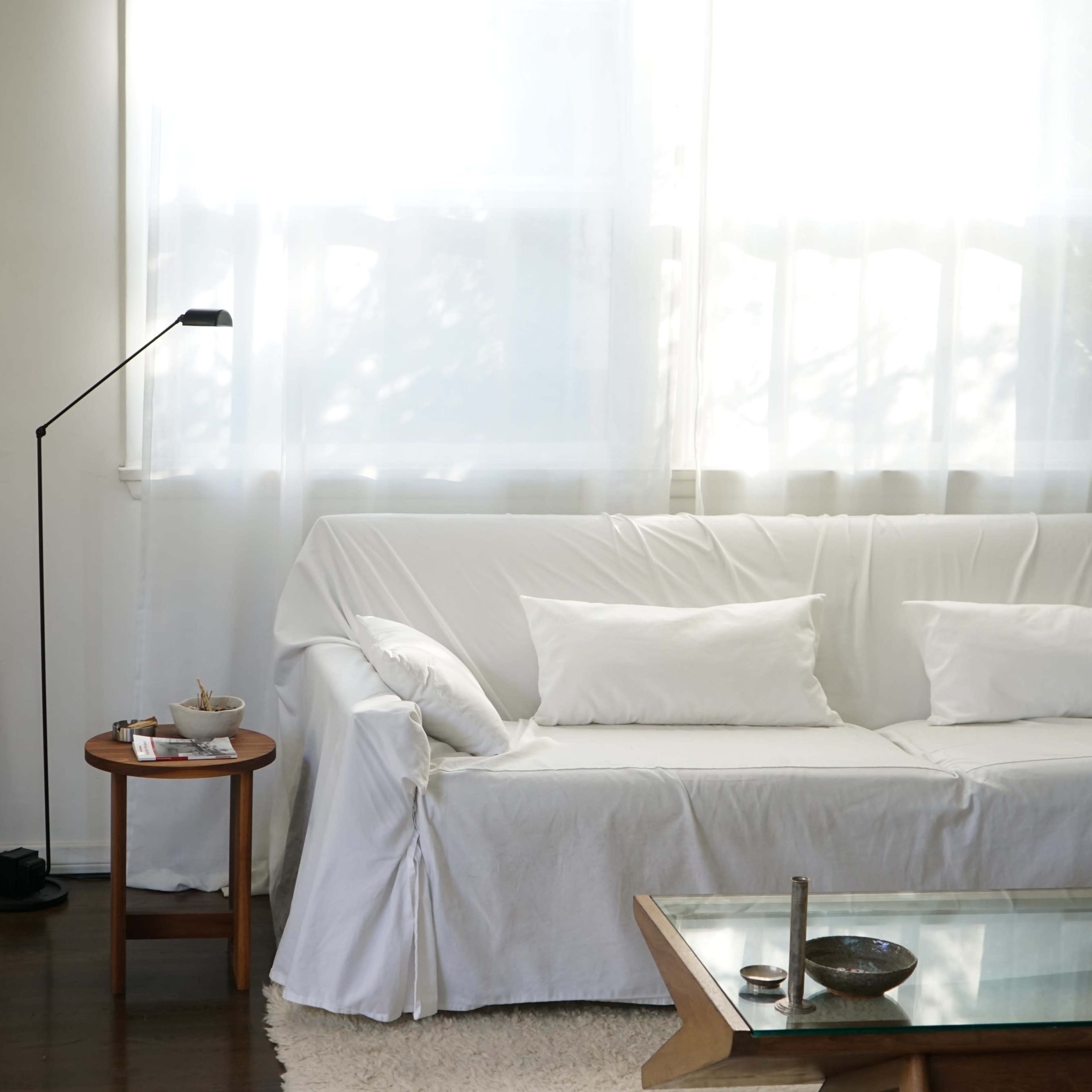 A minimalist living room with a white sofa covered in a slipcover, a wooden side table, a floor lamp, and natural light streaming through sheer curtains.