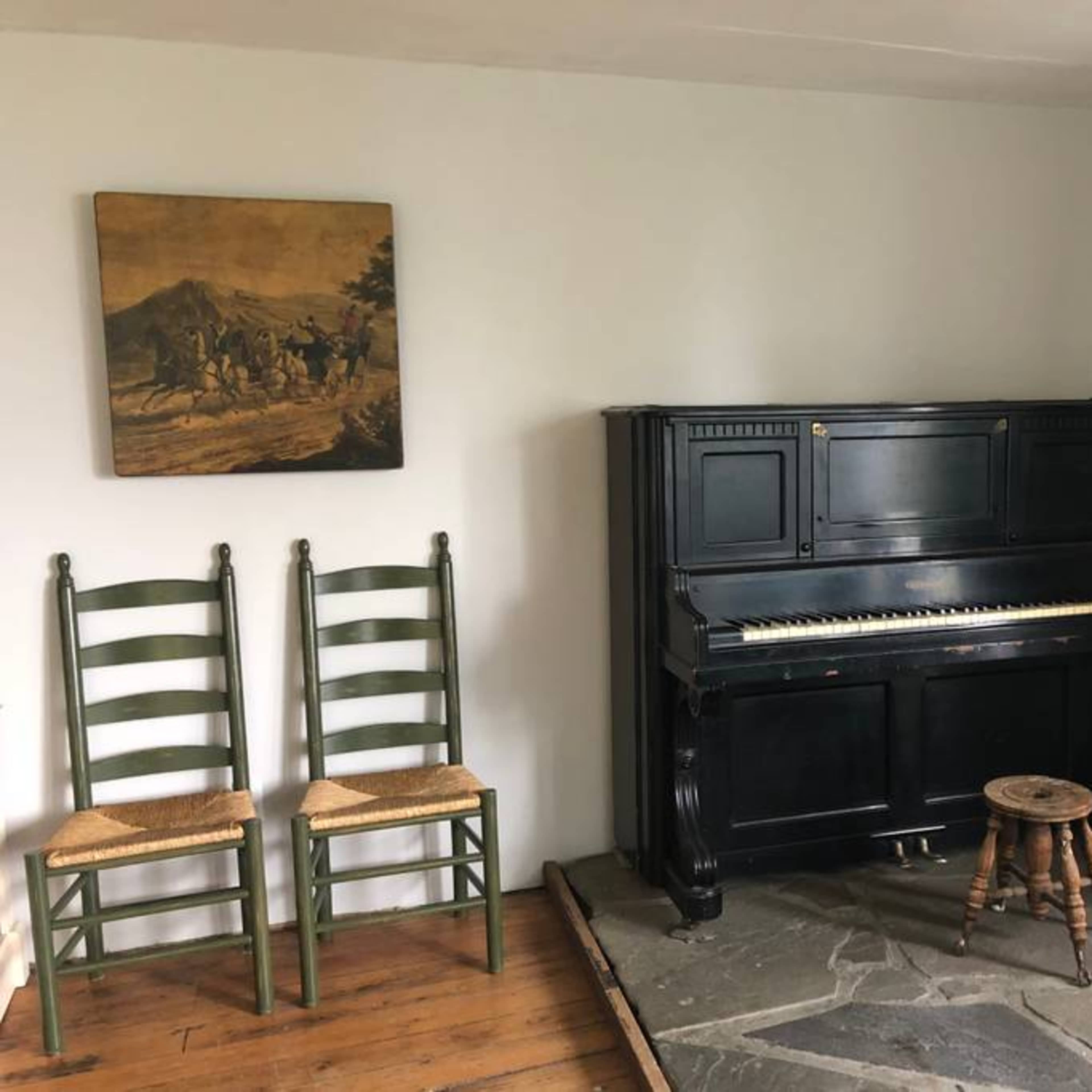 A room with a black upright piano, a small stool, two green wooden chairs, and a banjo hanging on the wall beside a painting of a group of people.
