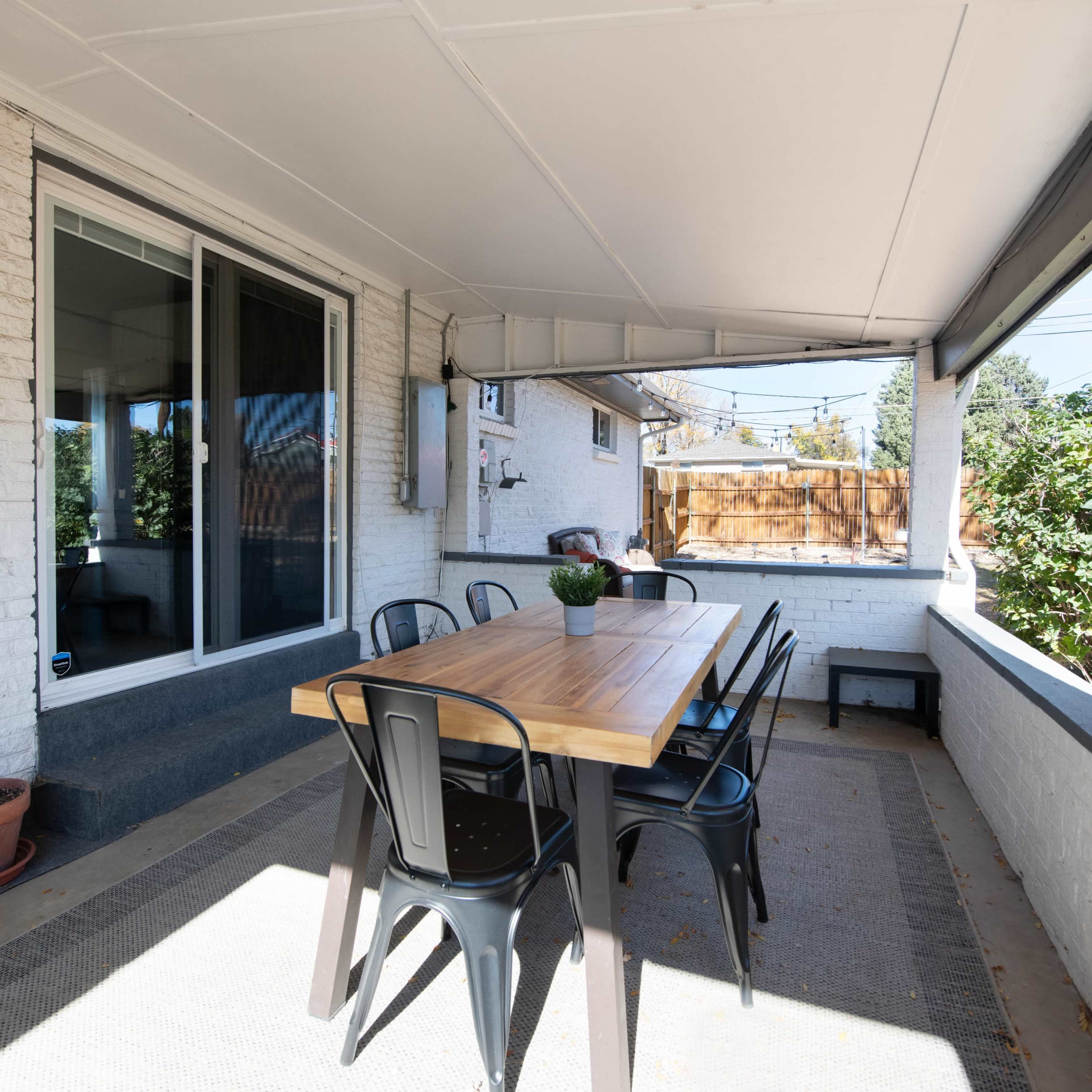 A wooden table with black chairs is set under a covered patio that features white brick walls and large windows.