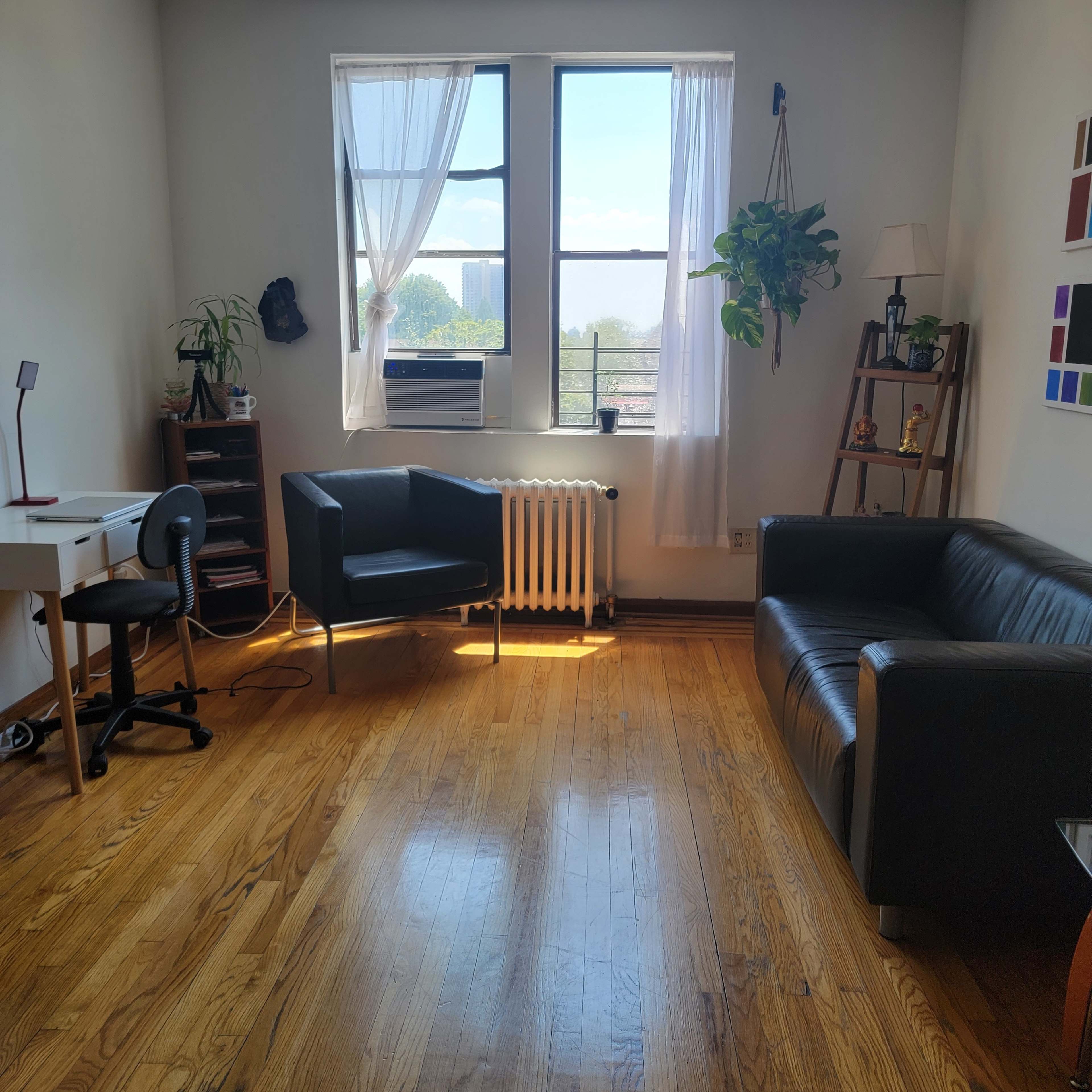A bright room with hardwood floors, featuring a desk and chair, two seating areas, and large windows with sheer curtains.