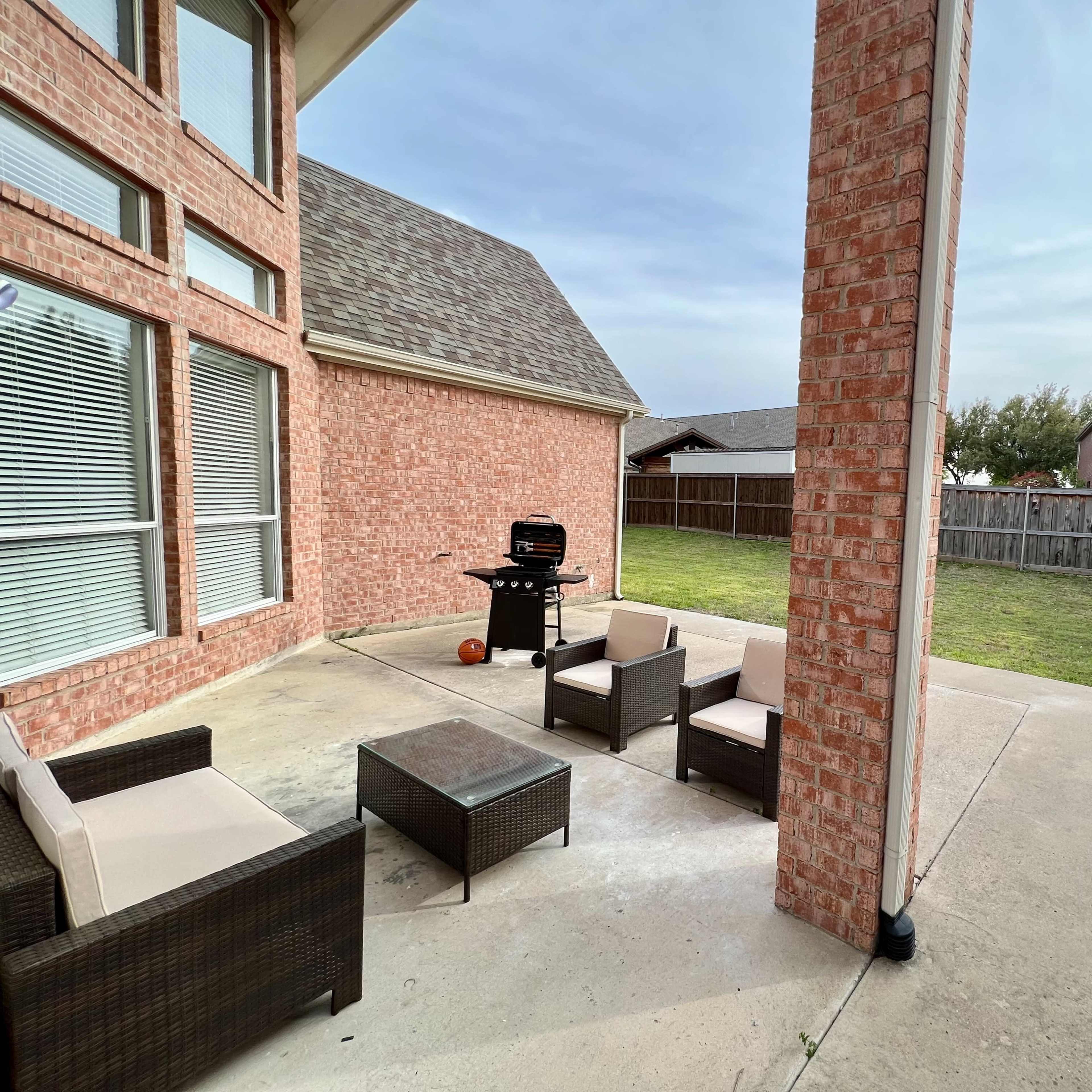 A brick patio area features a barbecue grill, outdoor seating, and a grassy yard enclosed by a fence.