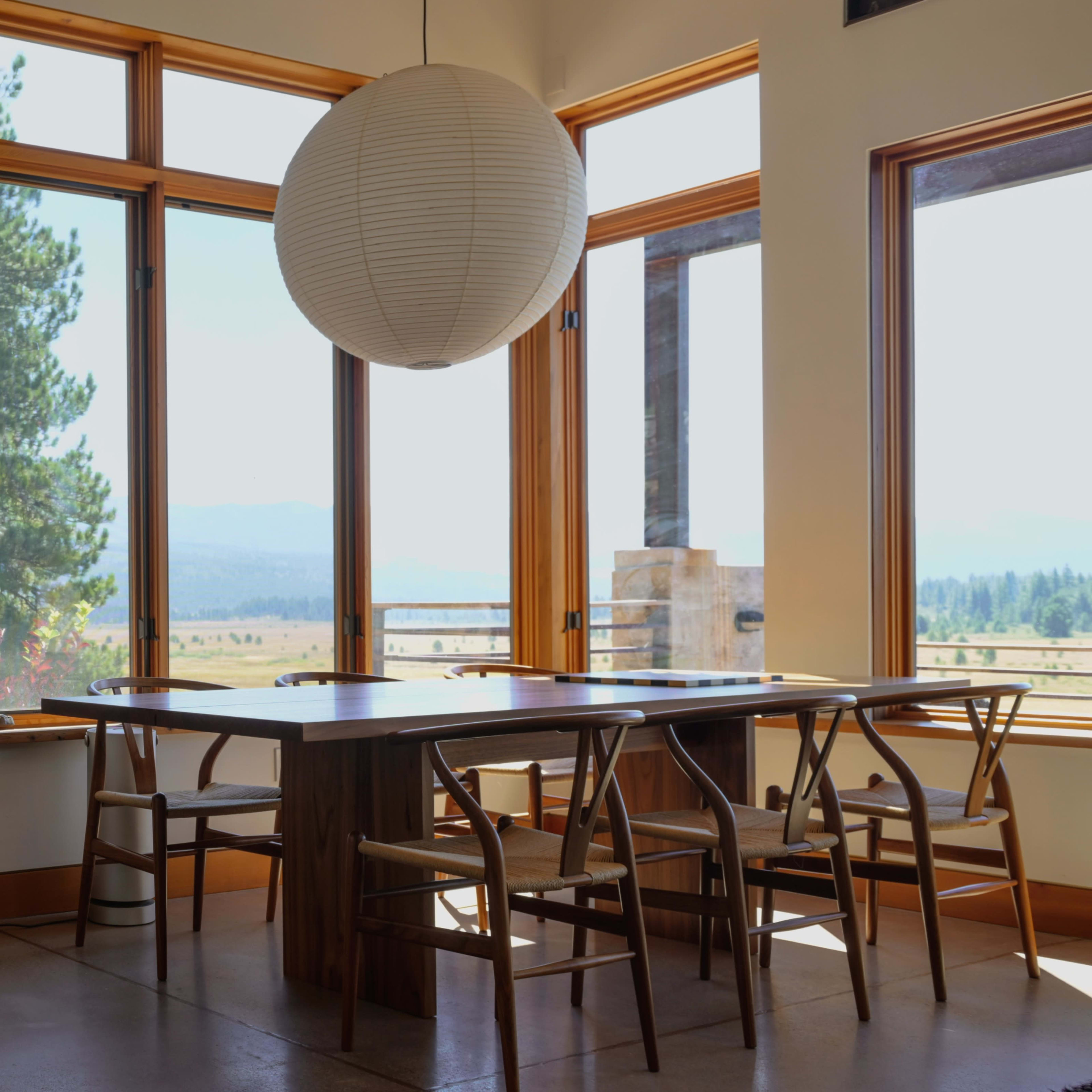 A modern dining area features a wooden table surrounded by chairs and a large pendant light, with large windows offering a view of the landscape.