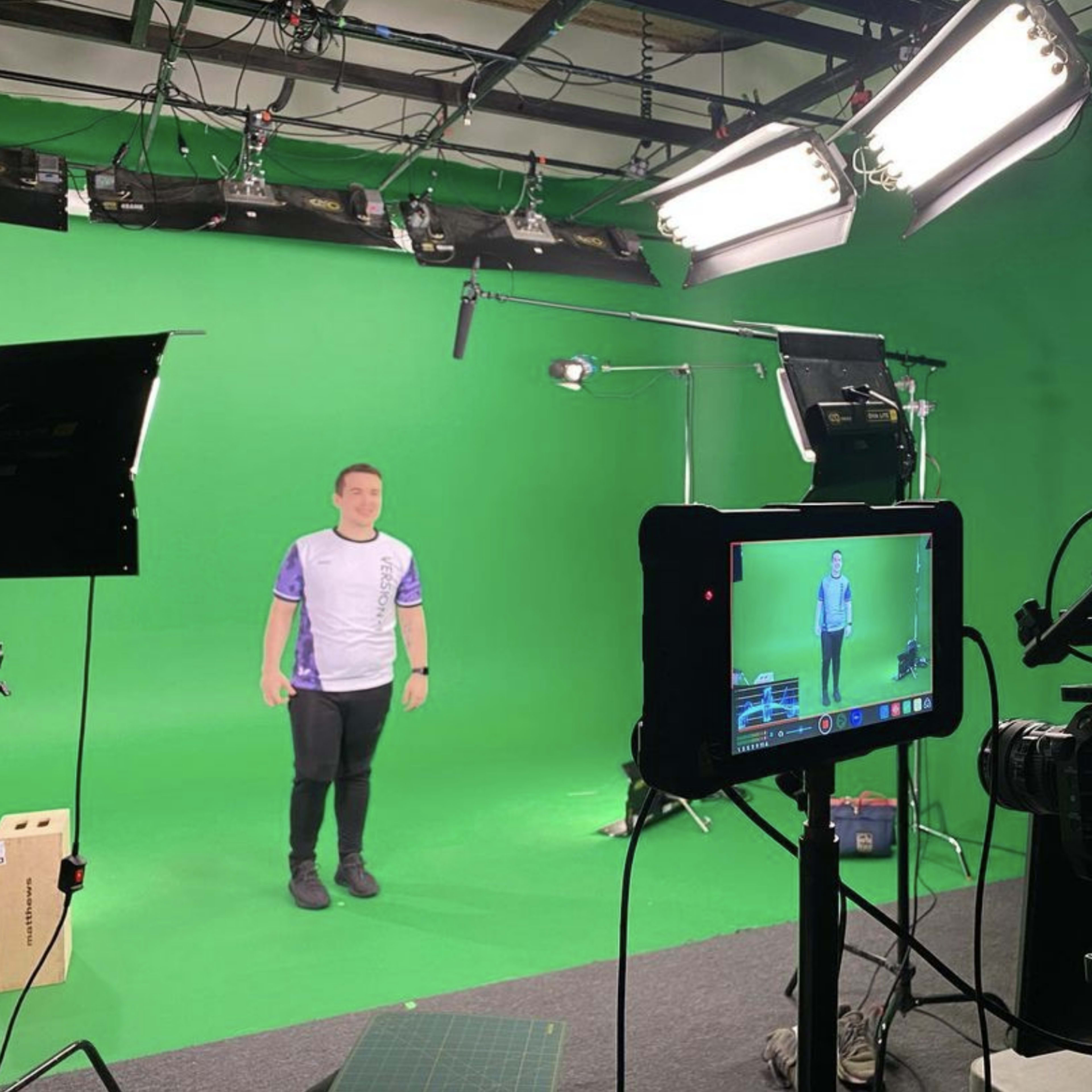 A person stands in front of a green screen in a video production studio, with camera equipment and lights positioned around the scene.