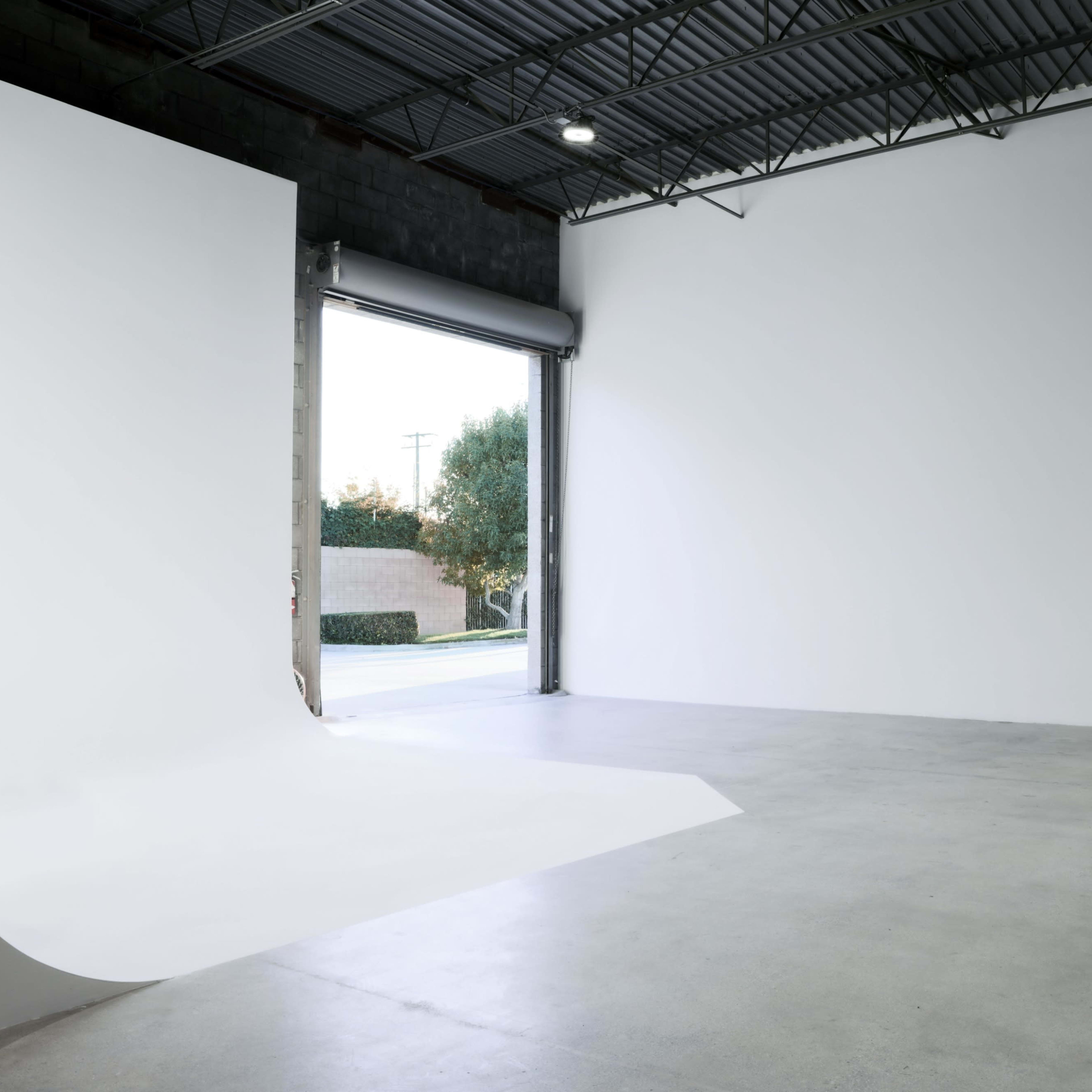 An empty indoor space features a large white backdrop and an open garage door leading to an outdoor area.