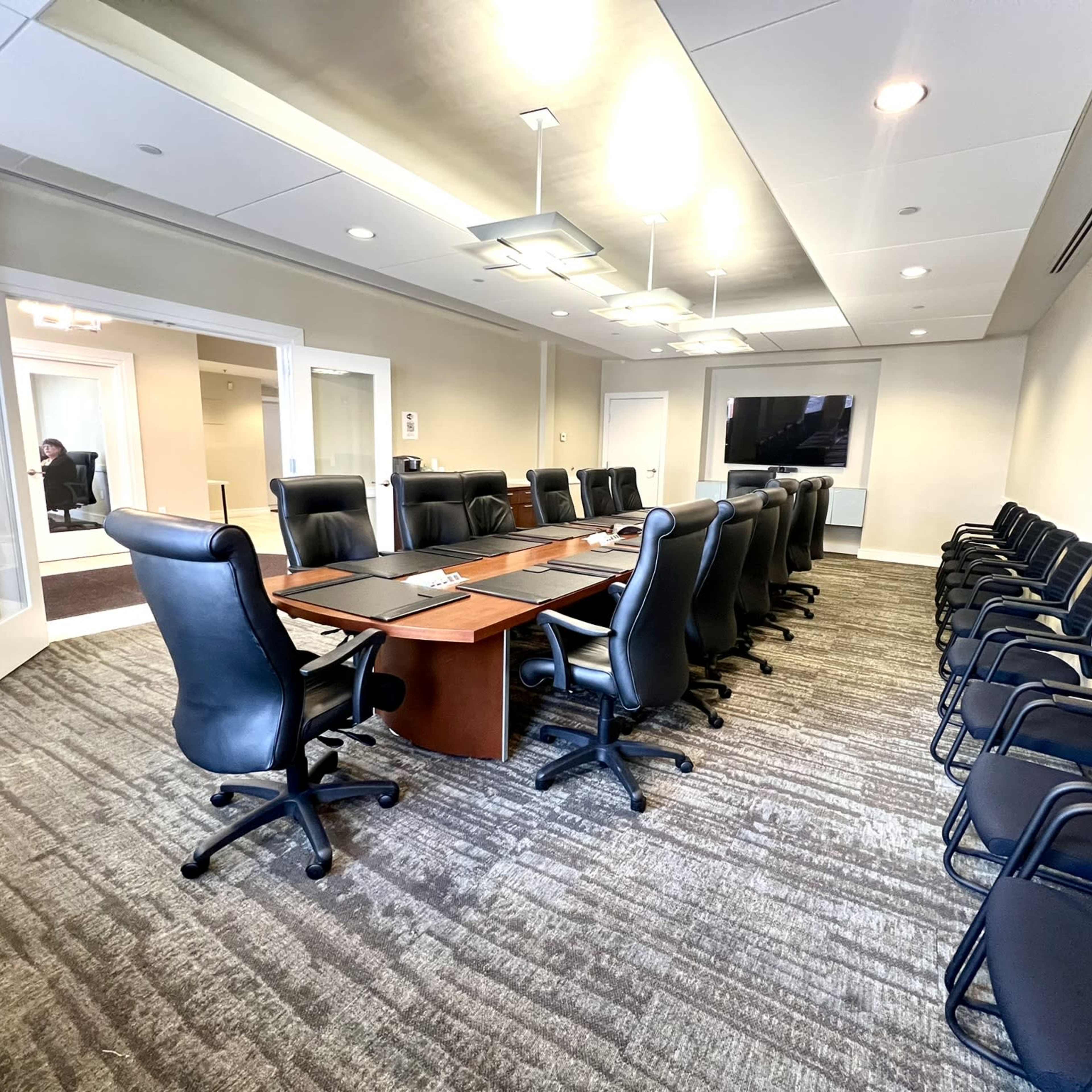 A large conference room features a long rectangular table surrounded by black leather chairs, with additional chairs aligned against the walls and a TV screen mounted at one end.