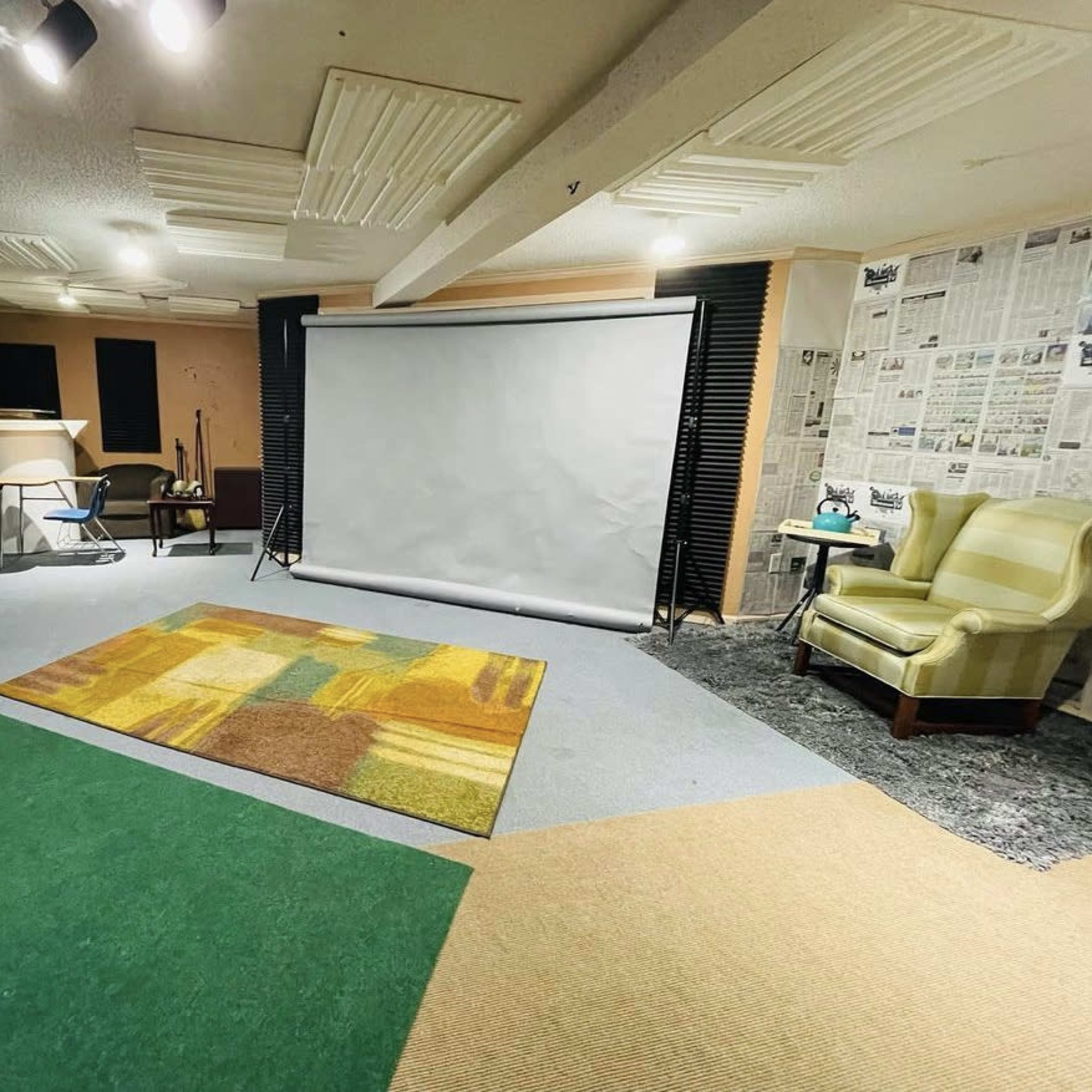 A basement room with a large projection screen, seating areas, and various floor coverings including green and beige carpet.