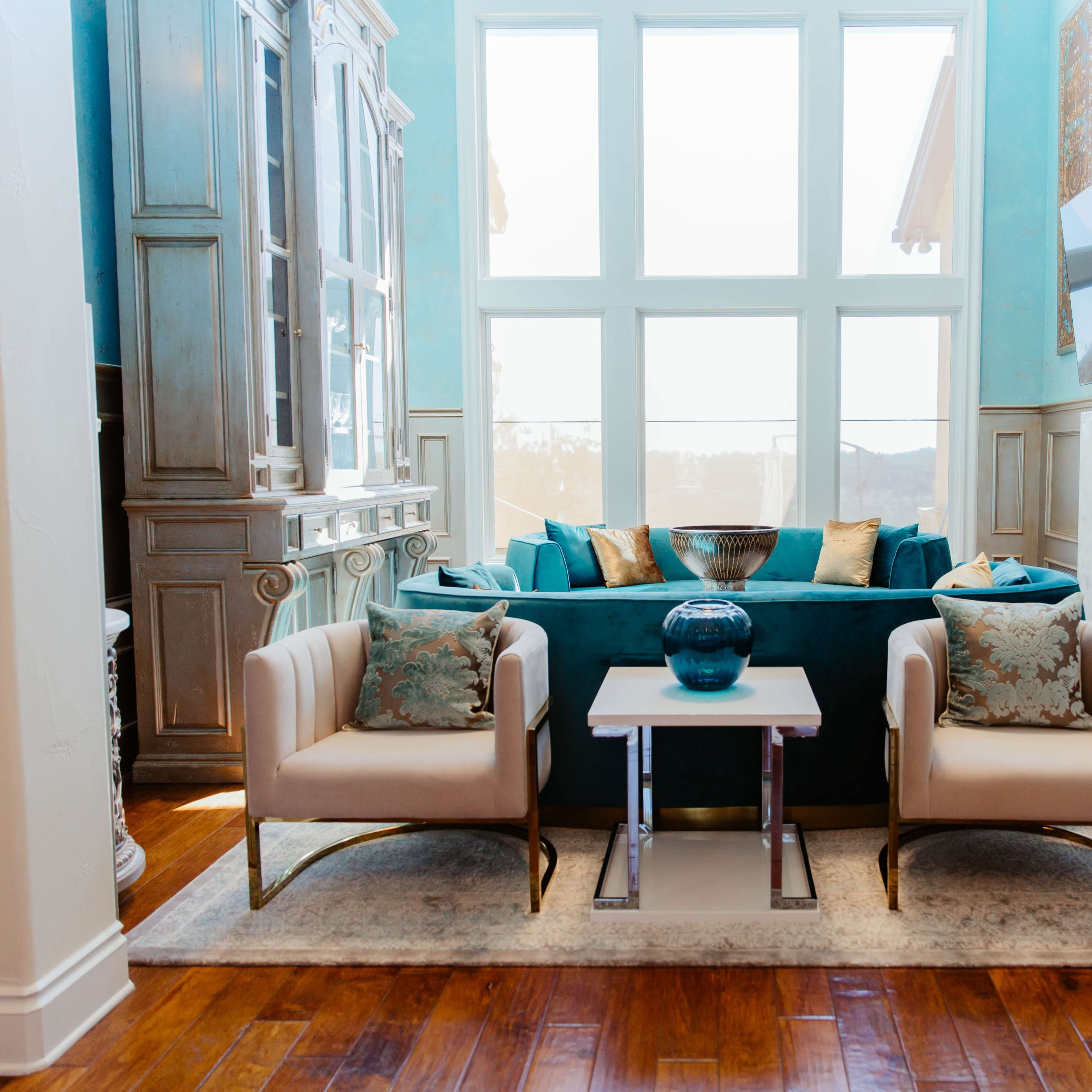 A modern living room featuring a turquoise couch, two white chairs, a glass coffee table, and large windows letting in natural light.