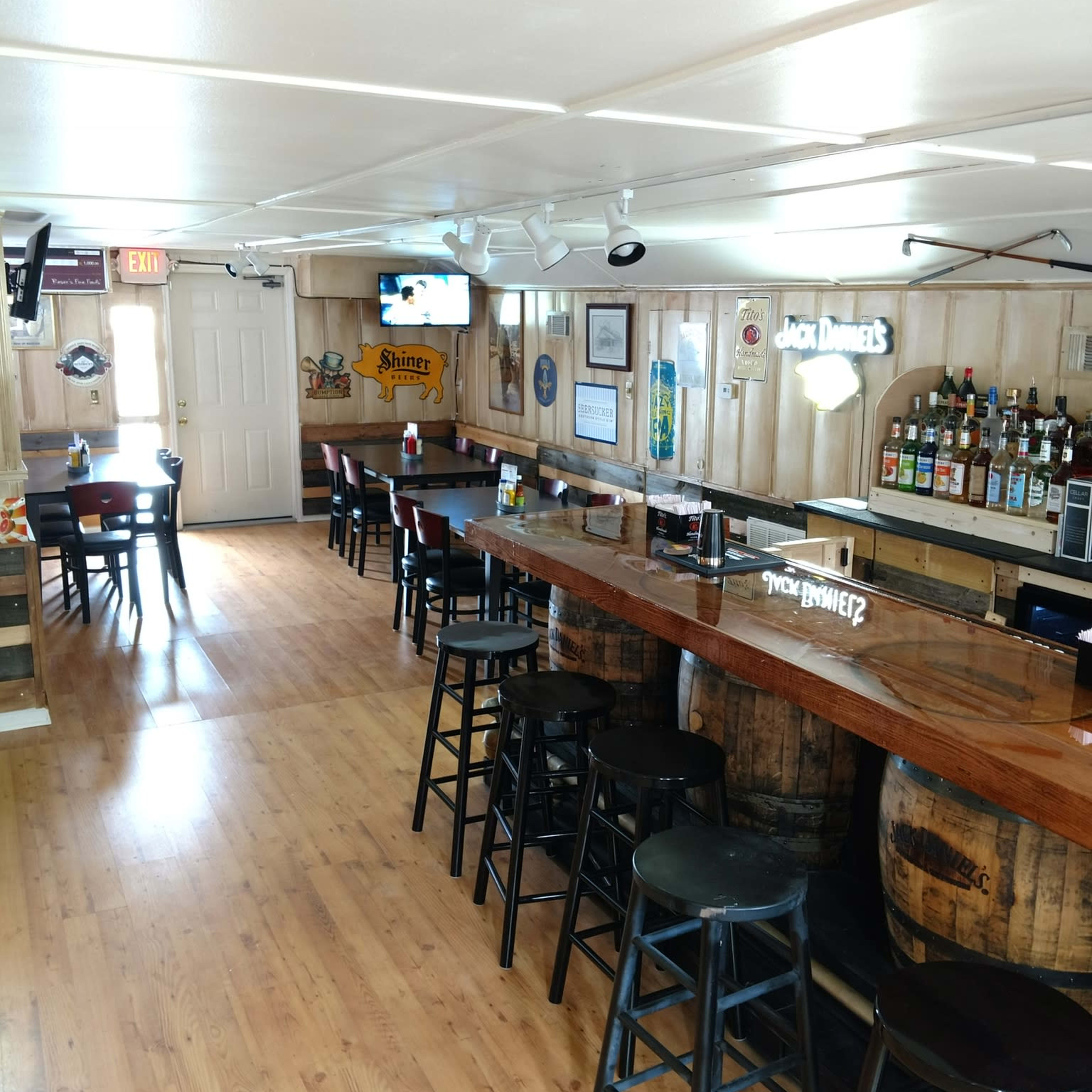 The interior of a rustic bar with wooden flooring, a long bar counter made from wood and barrels, and a few empty stools.