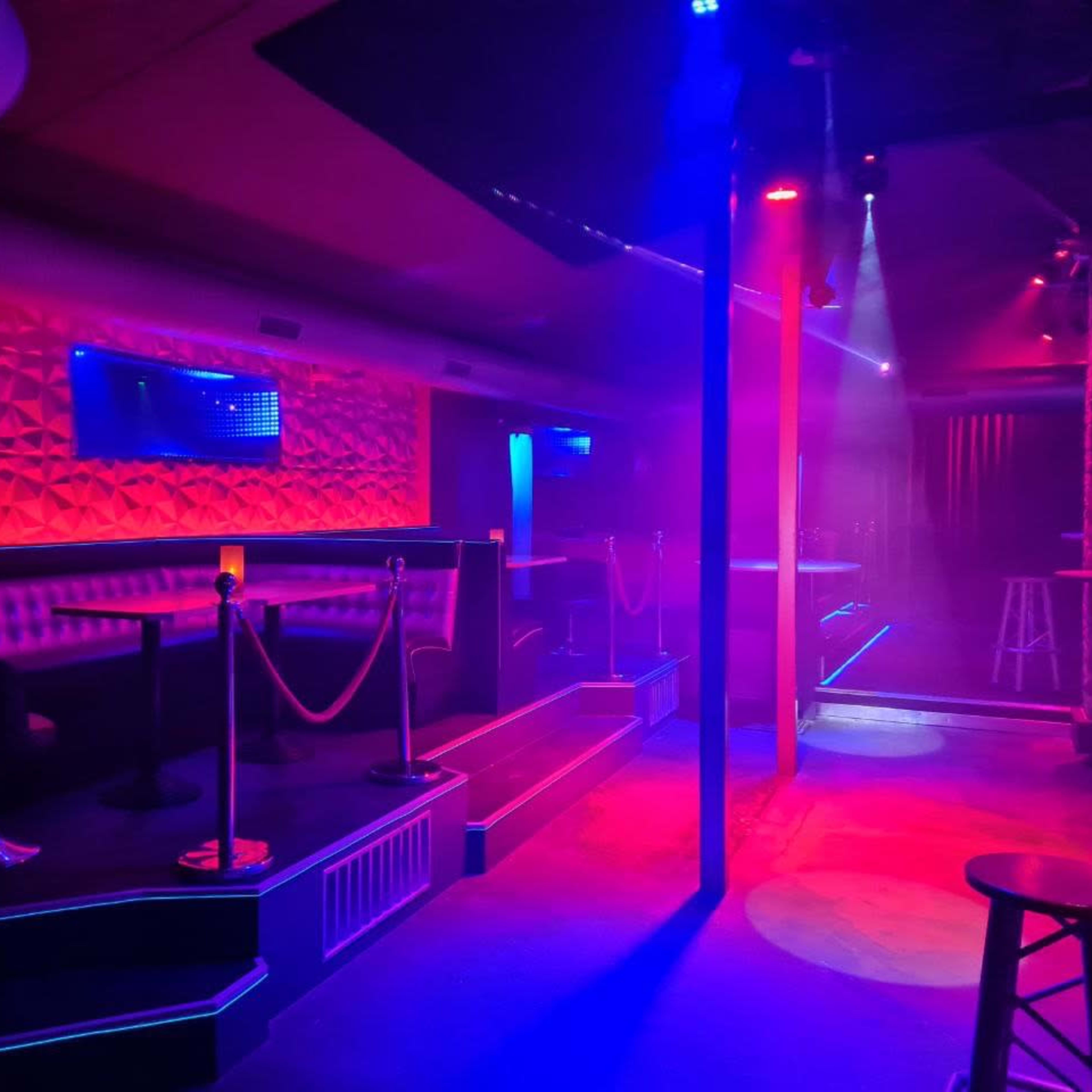 A dimly lit bar interior with blue and red lights, featuring seating areas separated by ropes and empty space illuminated by stage lights.
