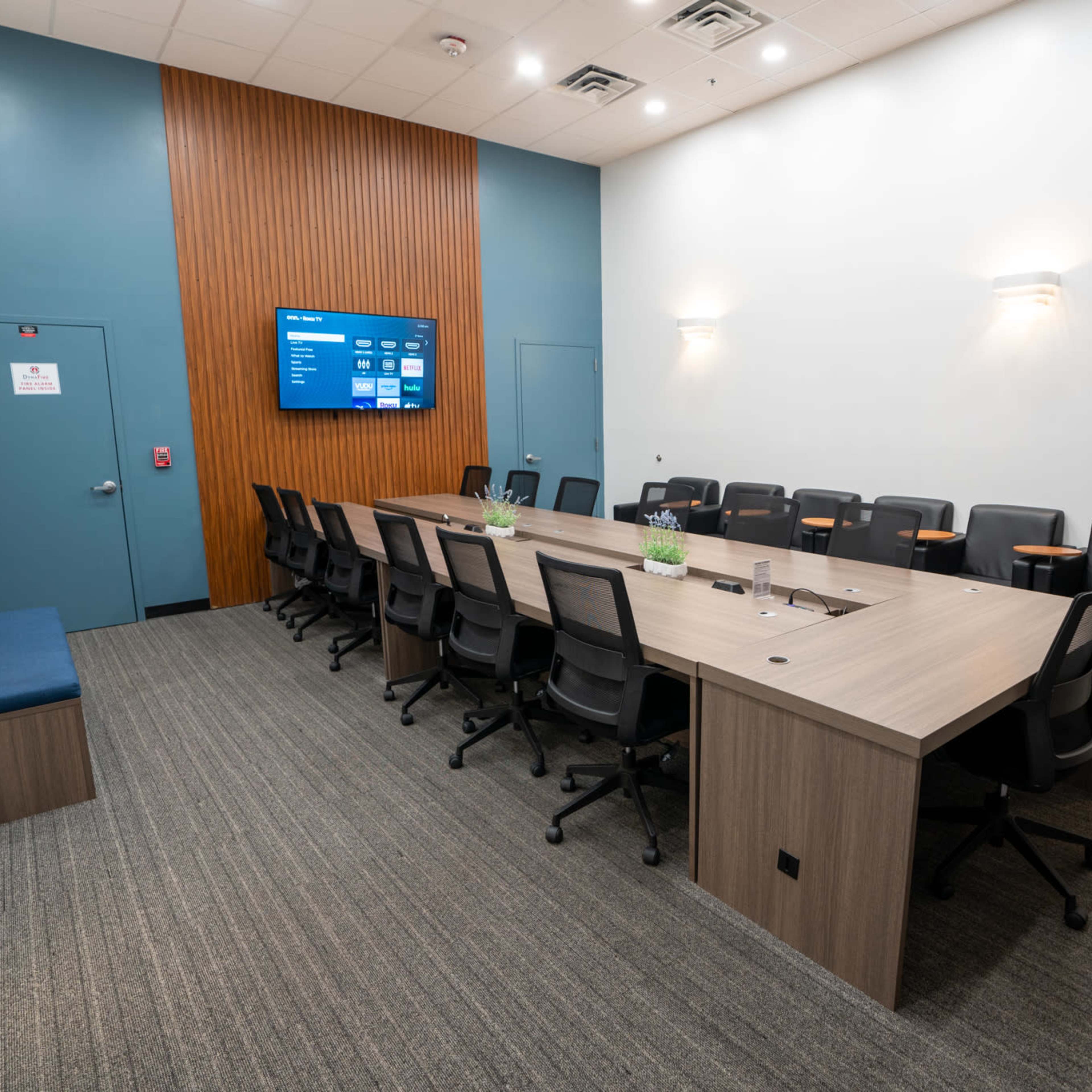 A modern conference room with a large table, multiple black chairs, and a wall-mounted screen.