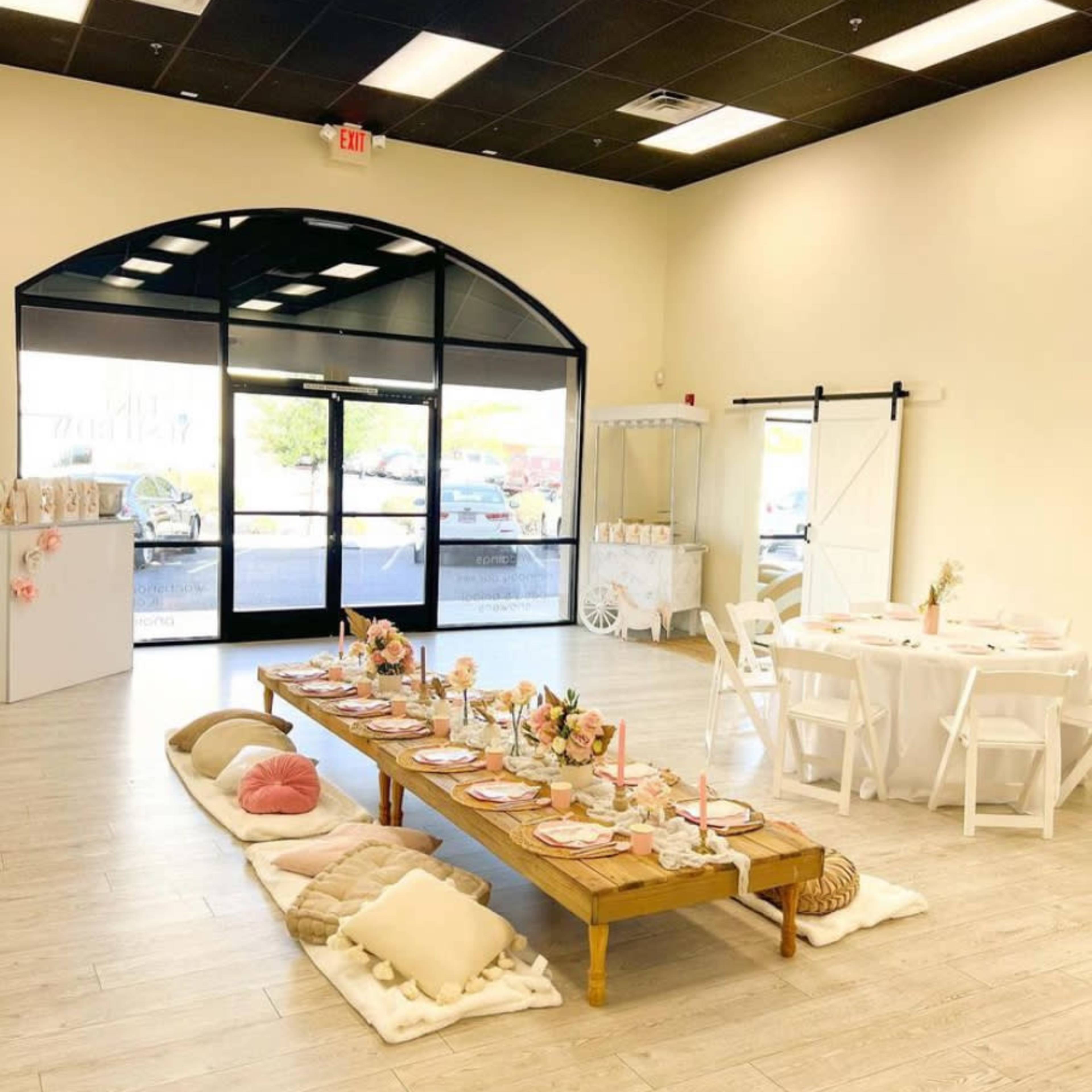An event space with a long wooden table set for dining, surrounded by white chairs and cushions on the floor, and large windows offering a view outside.