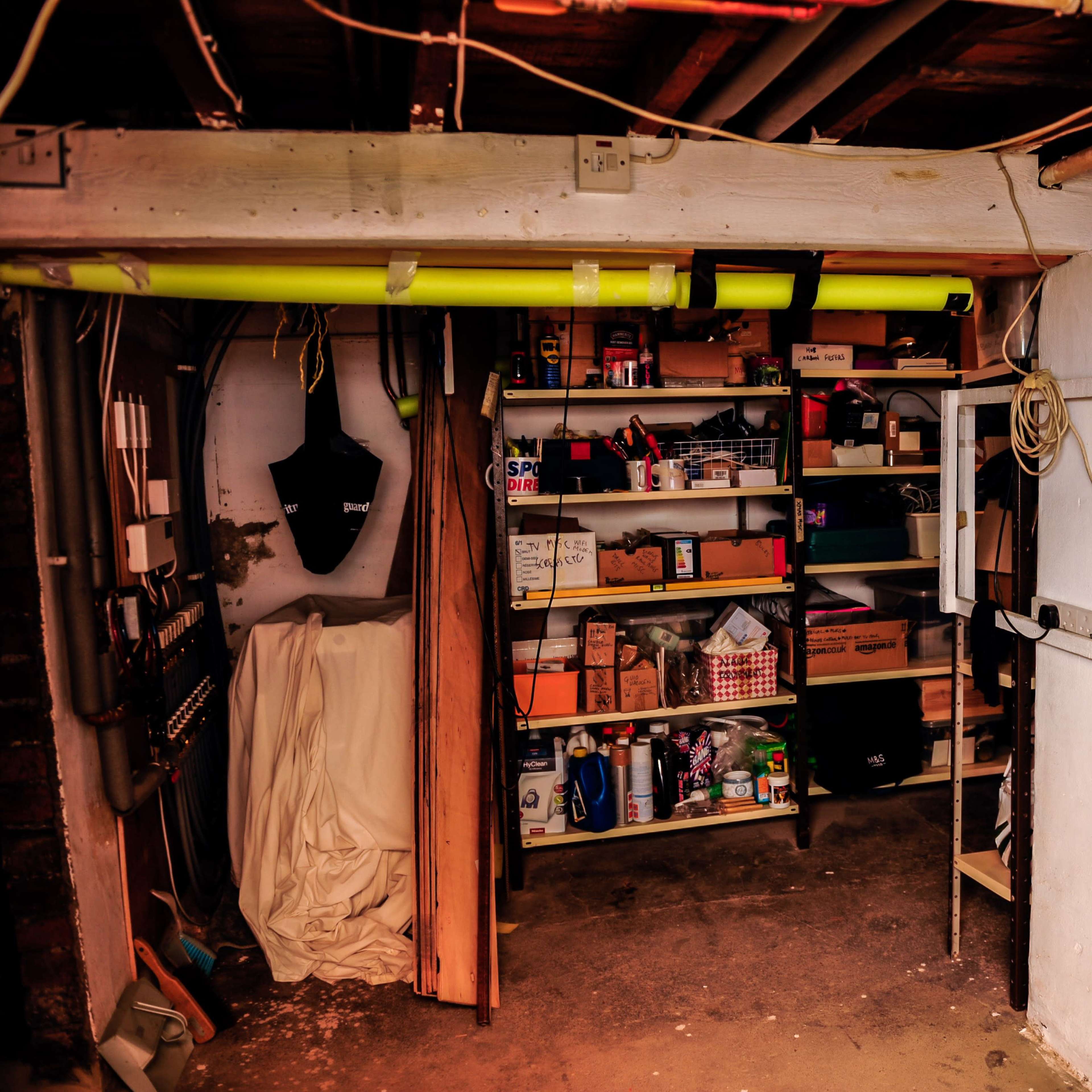 A dimly lit garage storage area features shelves filled with various boxes and containers, along with tools and materials along the walls.