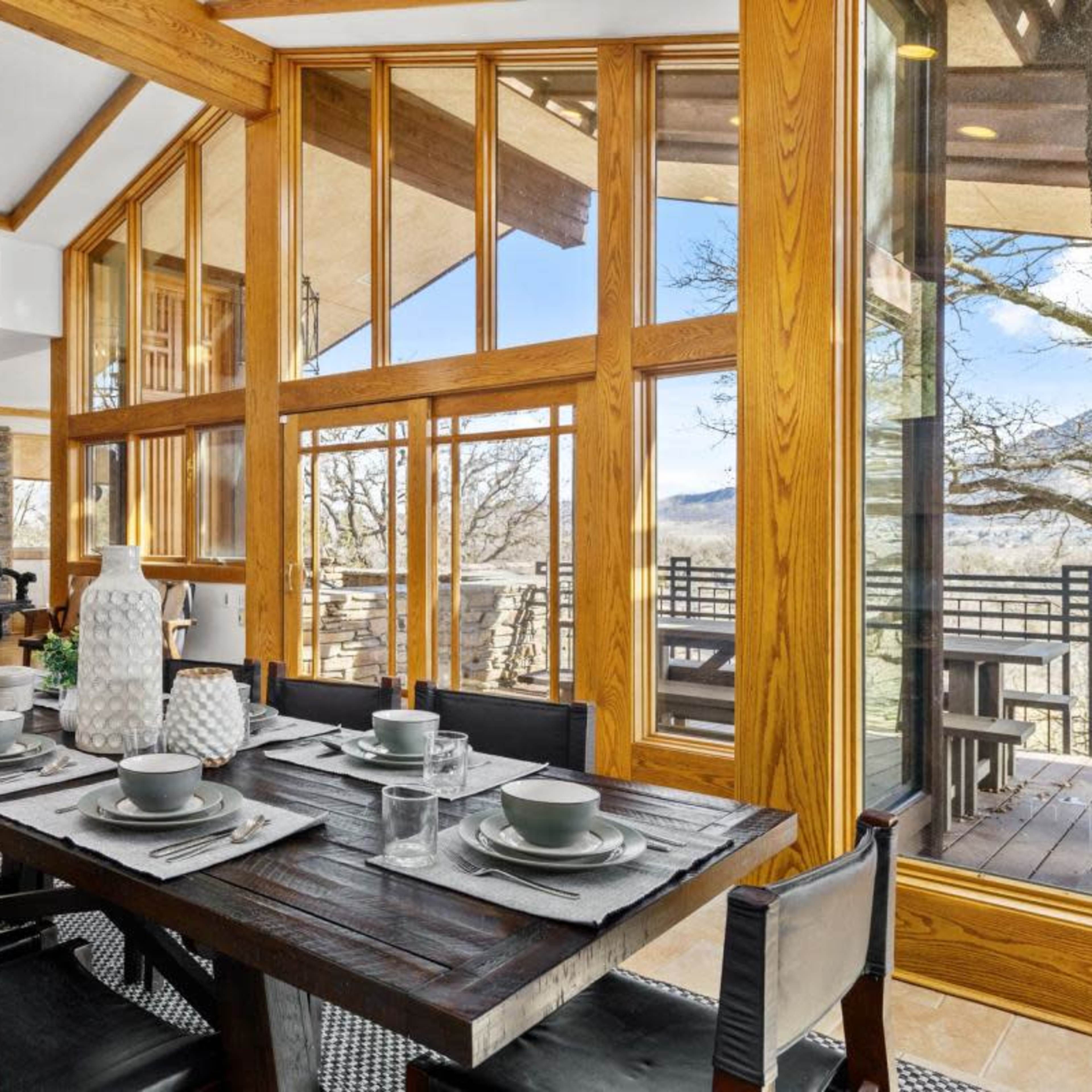 A modern dining area features a large wooden table set with dishes, surrounded by windows that provide a view of the outdoors.