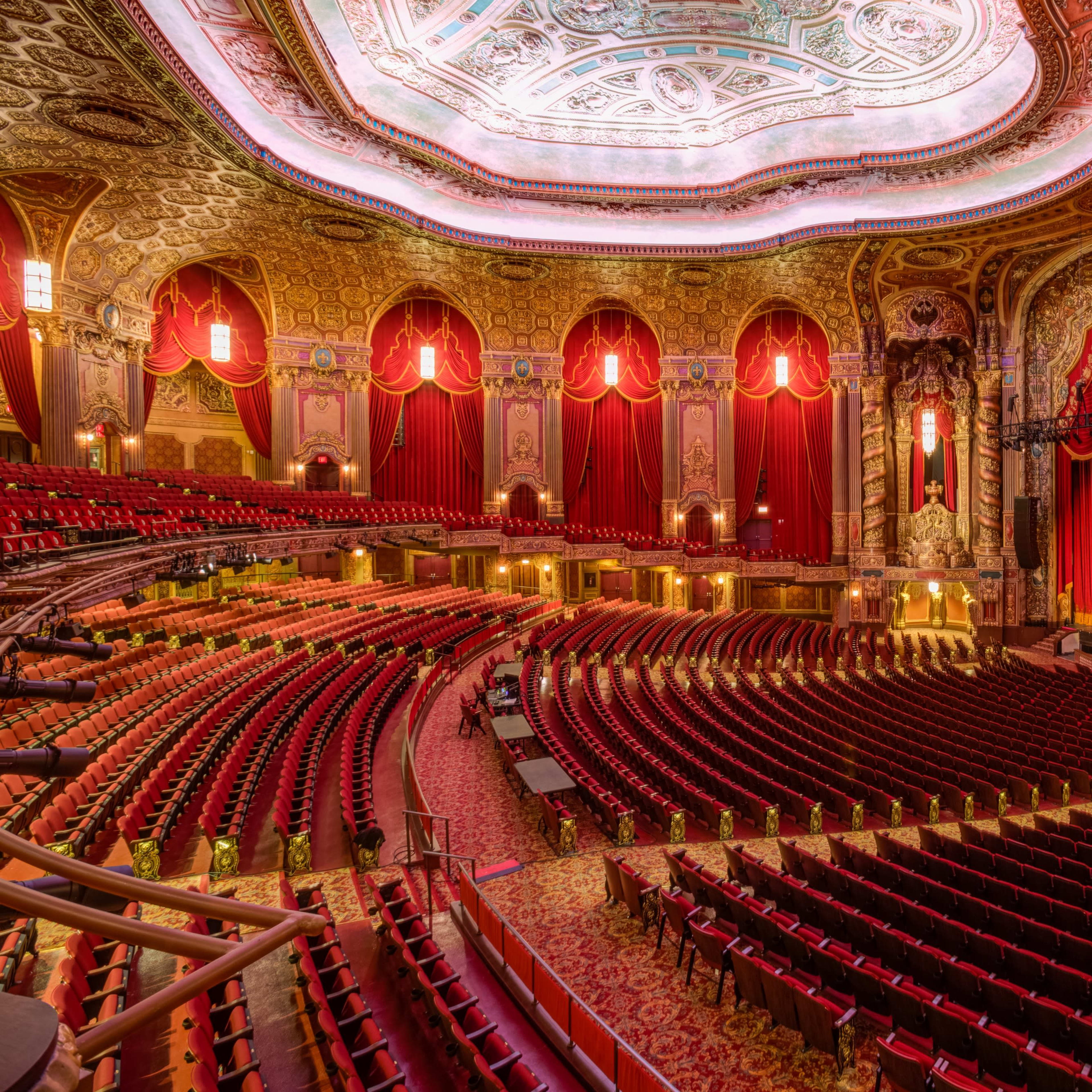 A grand theater interior with ornate decorations, red velvet curtains, and rows of empty seats facing the stage.
