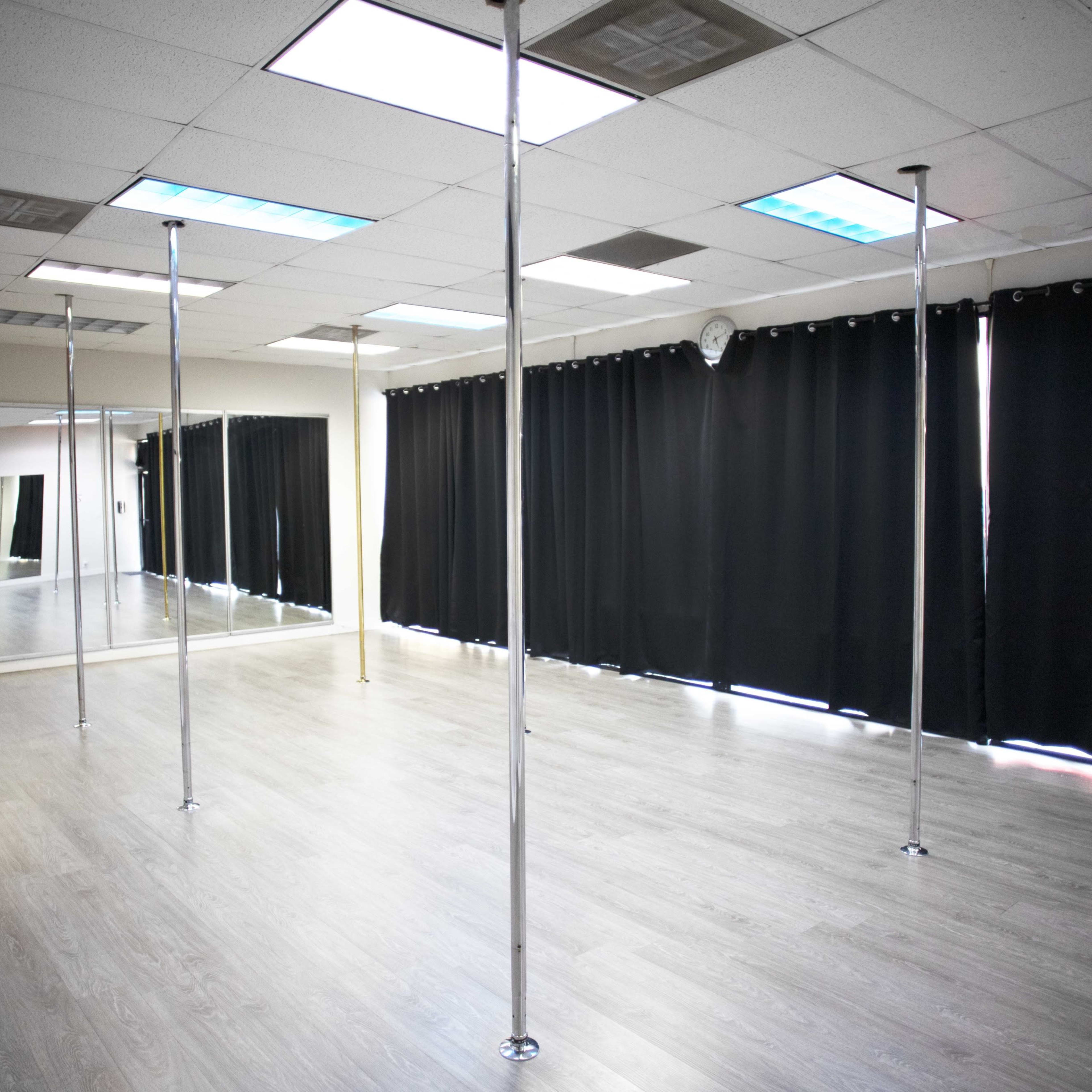 An empty dance studio with several vertical poles and large mirrors, illuminated by overhead lights and partially covered windows.