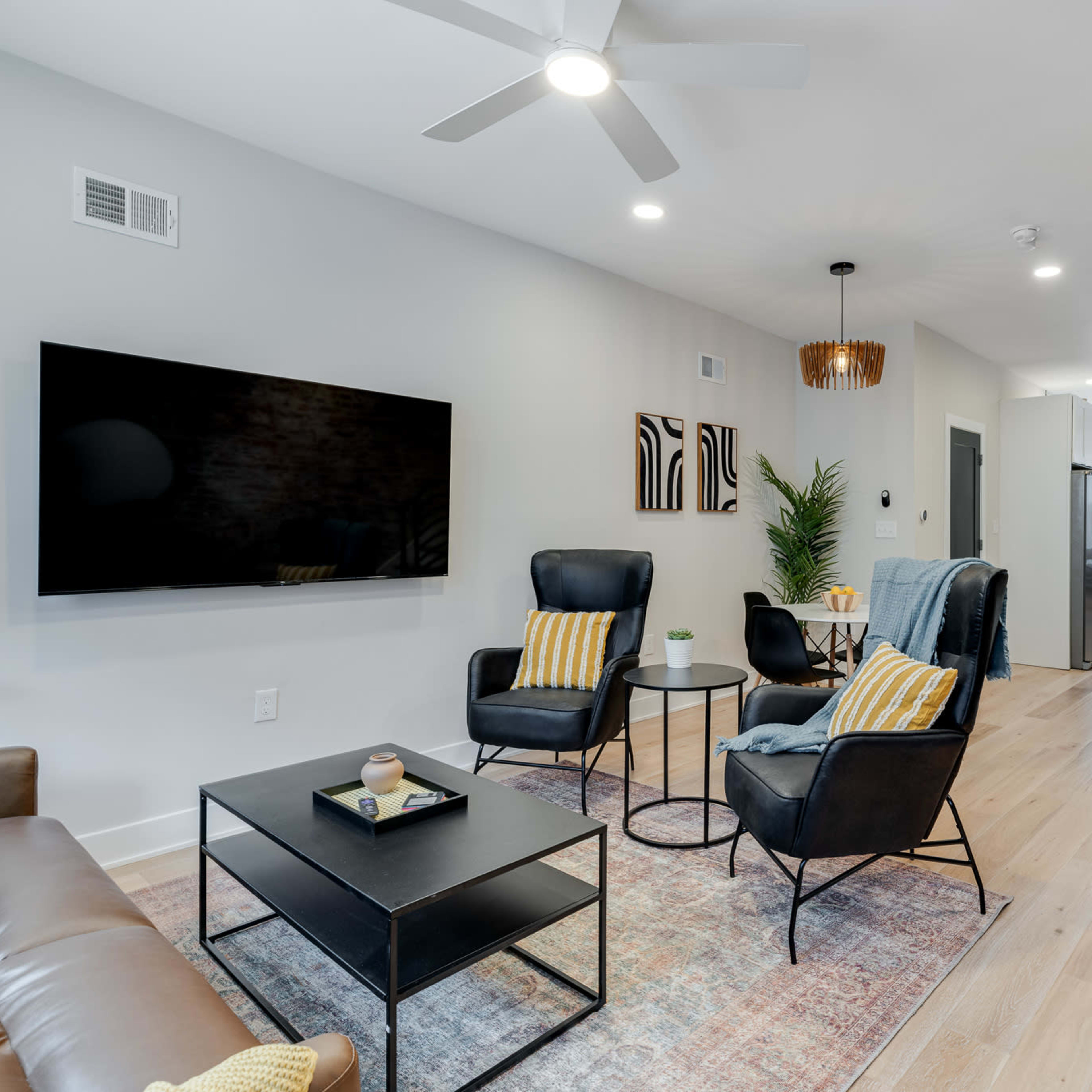 A modern living room features a TV mounted on the wall, two black chairs with yellow-striped cushions, a coffee table, and an open staircase leading to the upper floor.