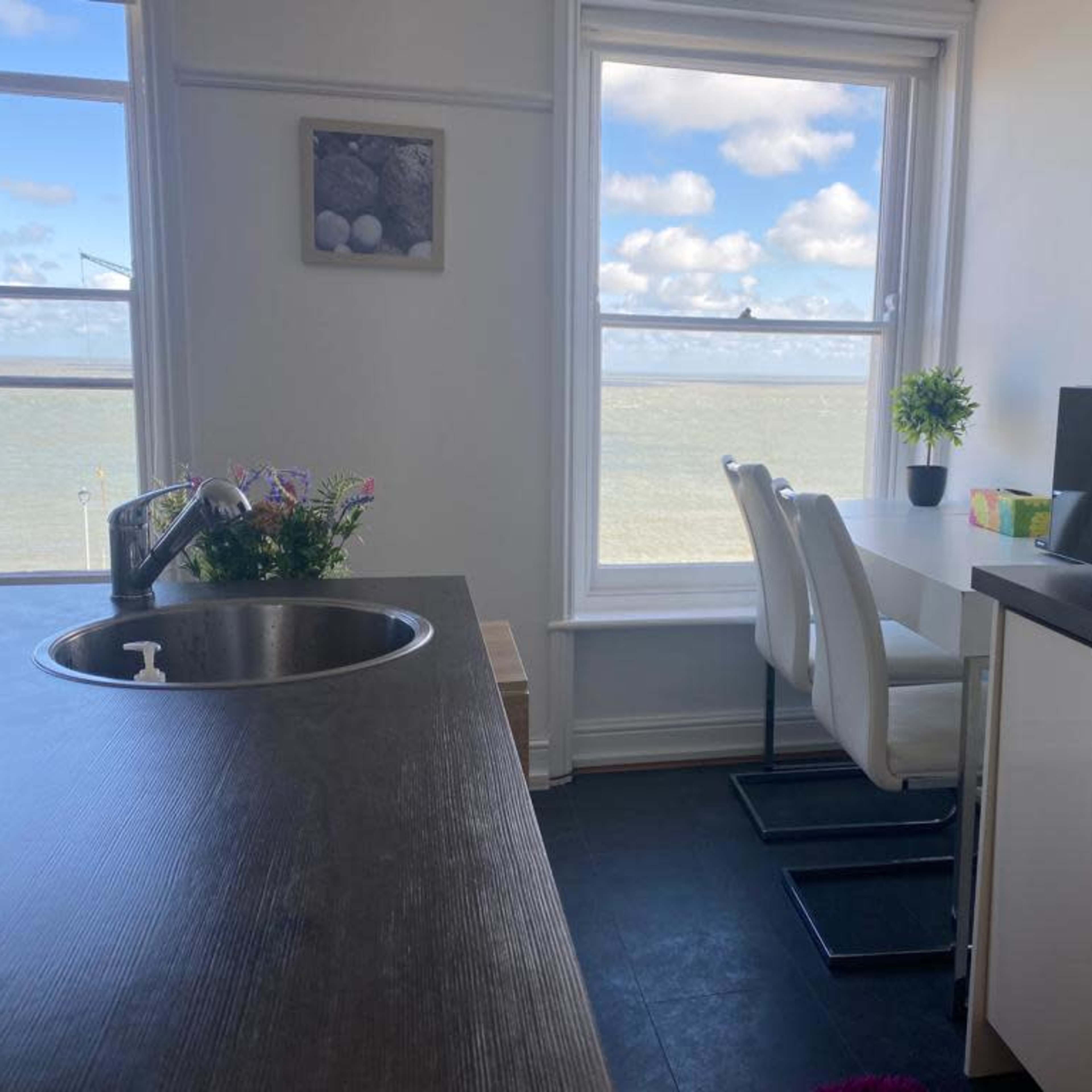 A modern kitchen space with a sink, a table with chairs, and a window offering a view of the sea and clouds.