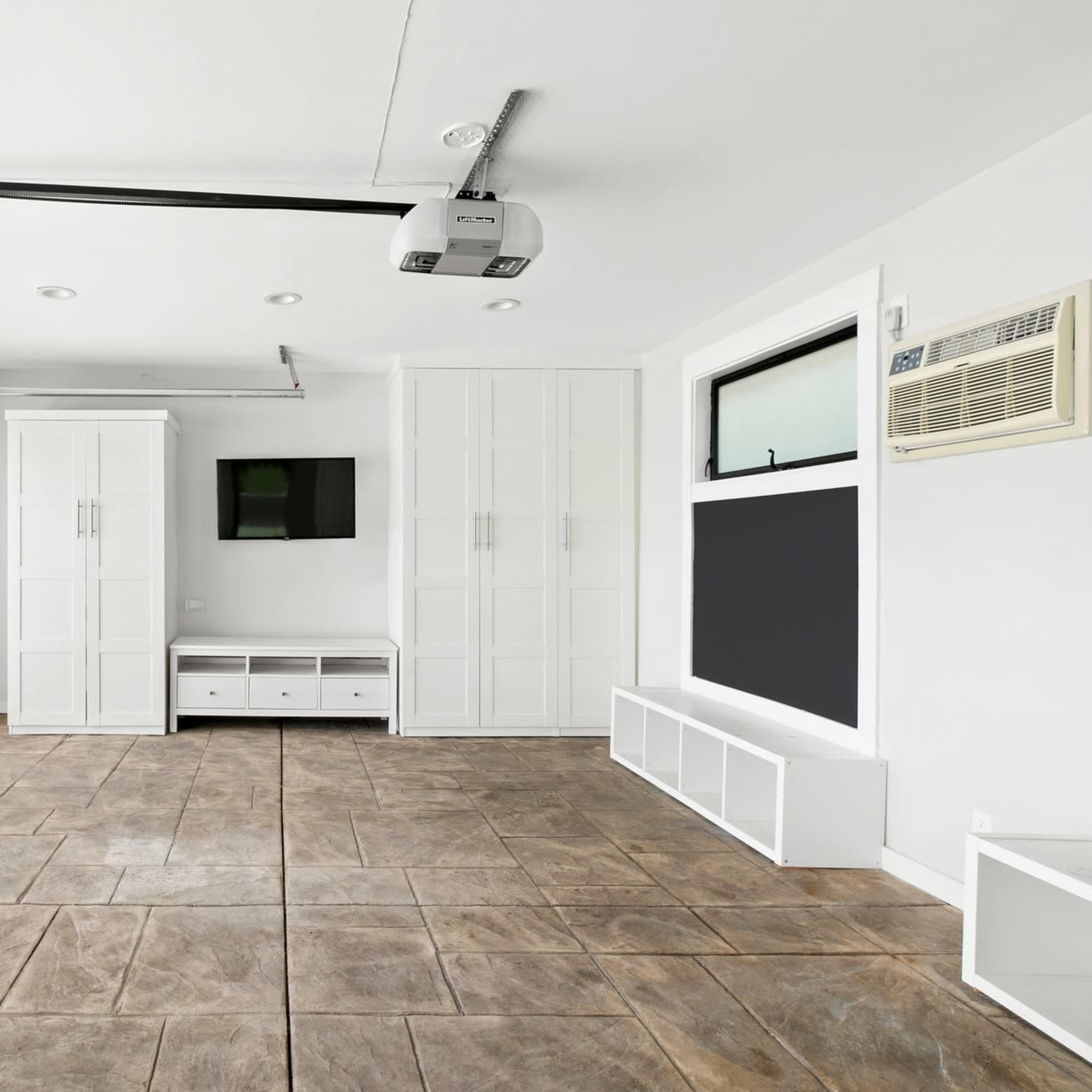 A modern garage with a large door, white cabinetry, a wall-mounted TV, and a concrete floor.