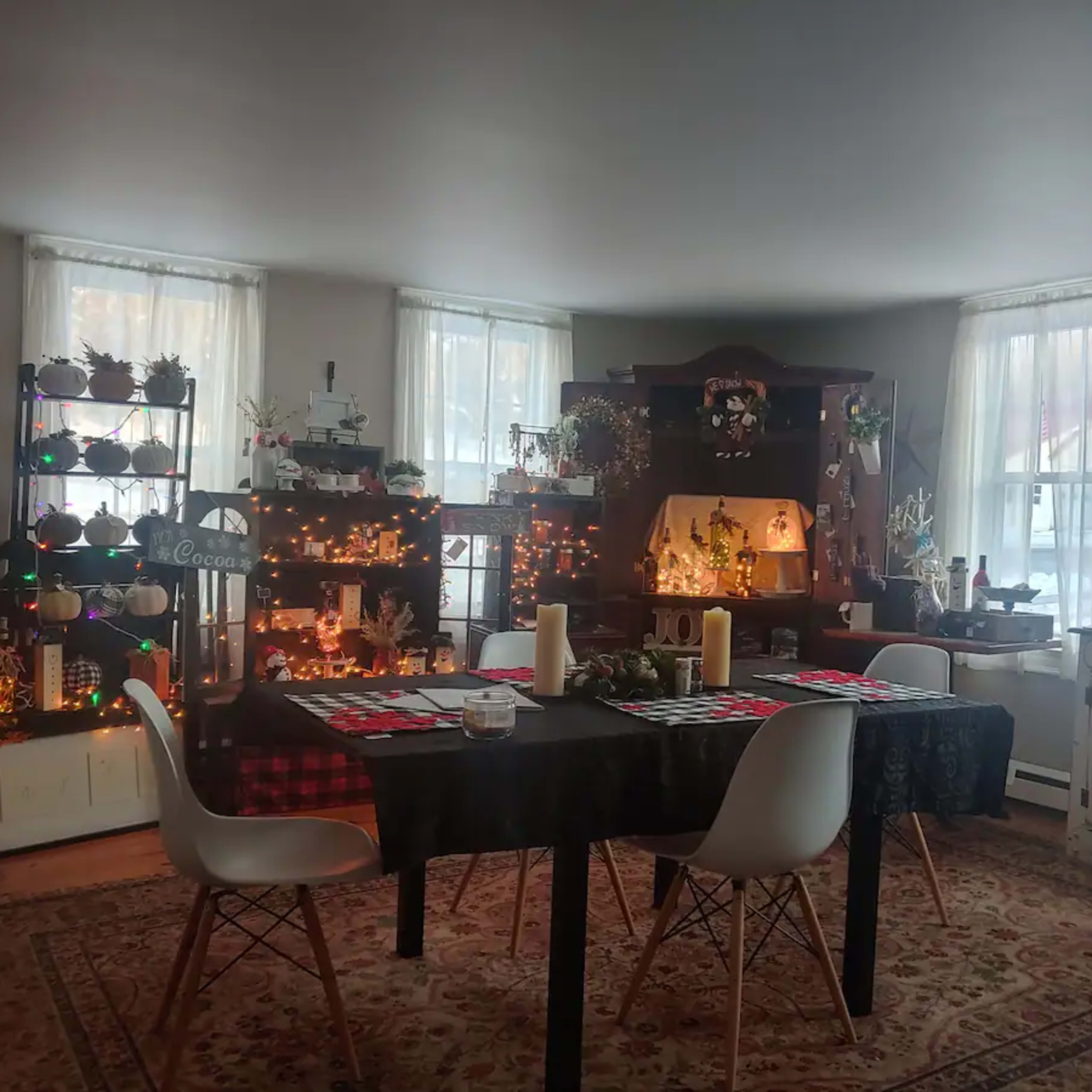 A dining area features a black table with white chairs, surrounded by decorative shelves filled with plants and lights, and large windows letting in natural light.