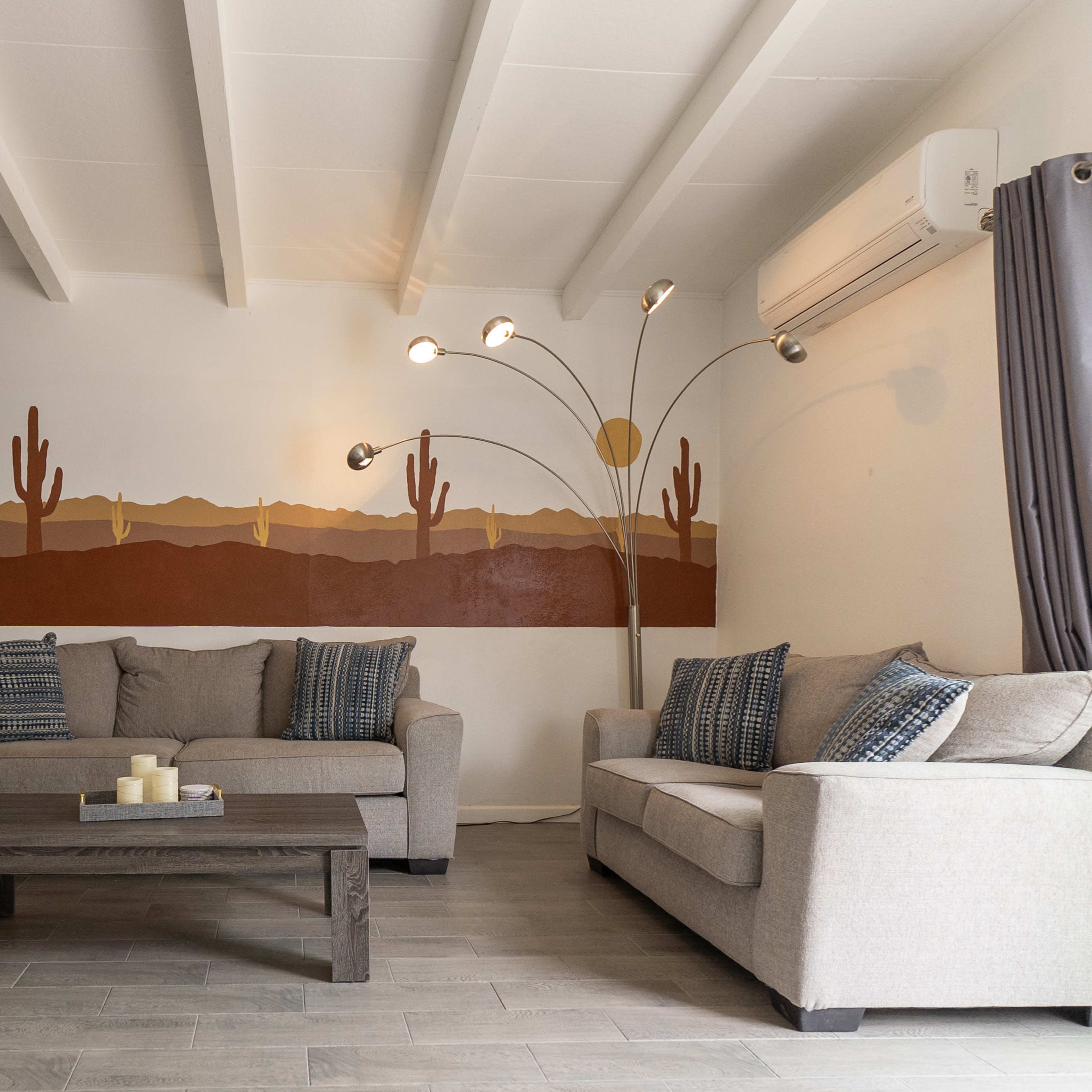 A modern living room features two light gray sofas, a wooden coffee table, and a decorative mural of cacti and mountains on the wall.