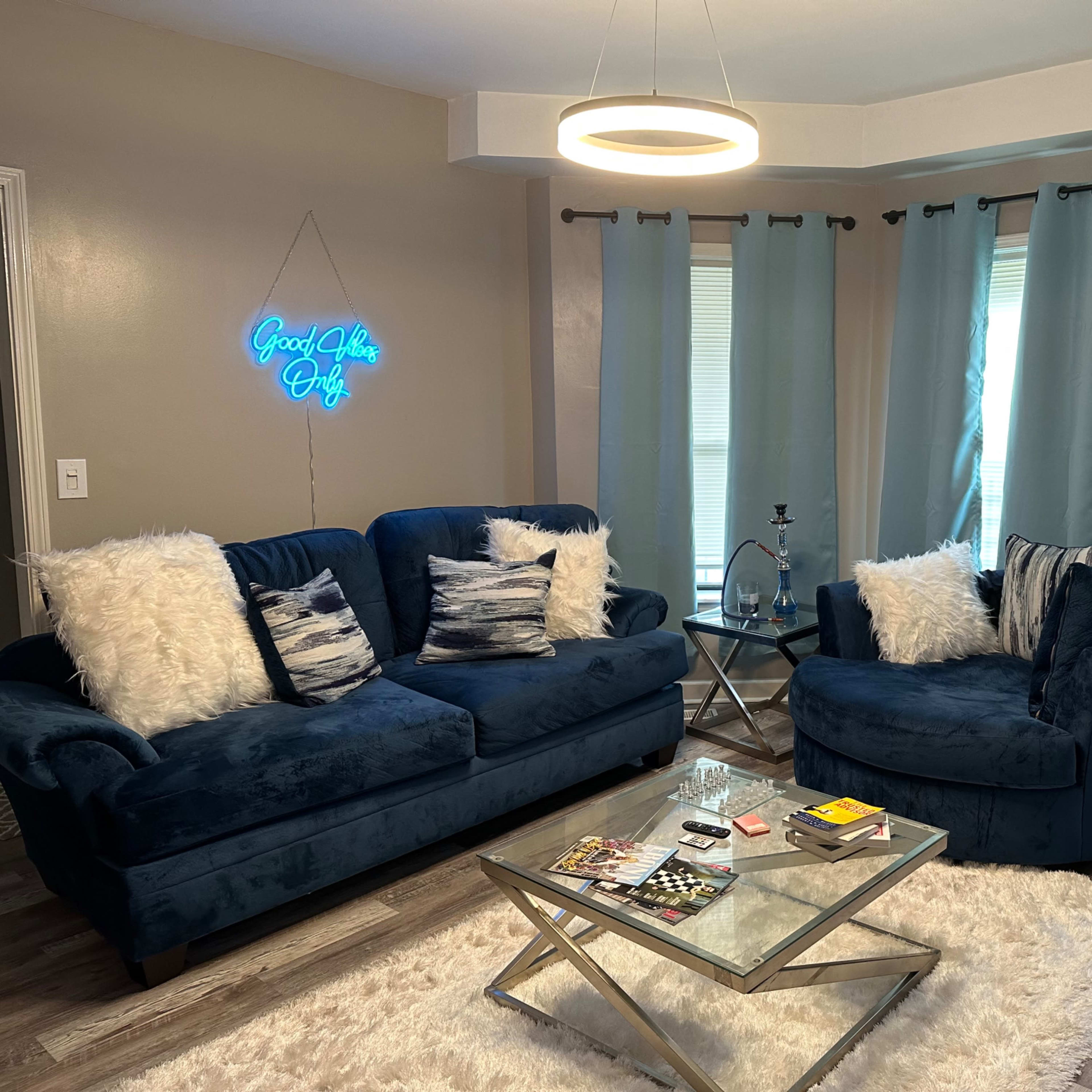 The living room features two blue sofas with decorative pillows, a glass coffee table, and large windows covered with light blue curtains.