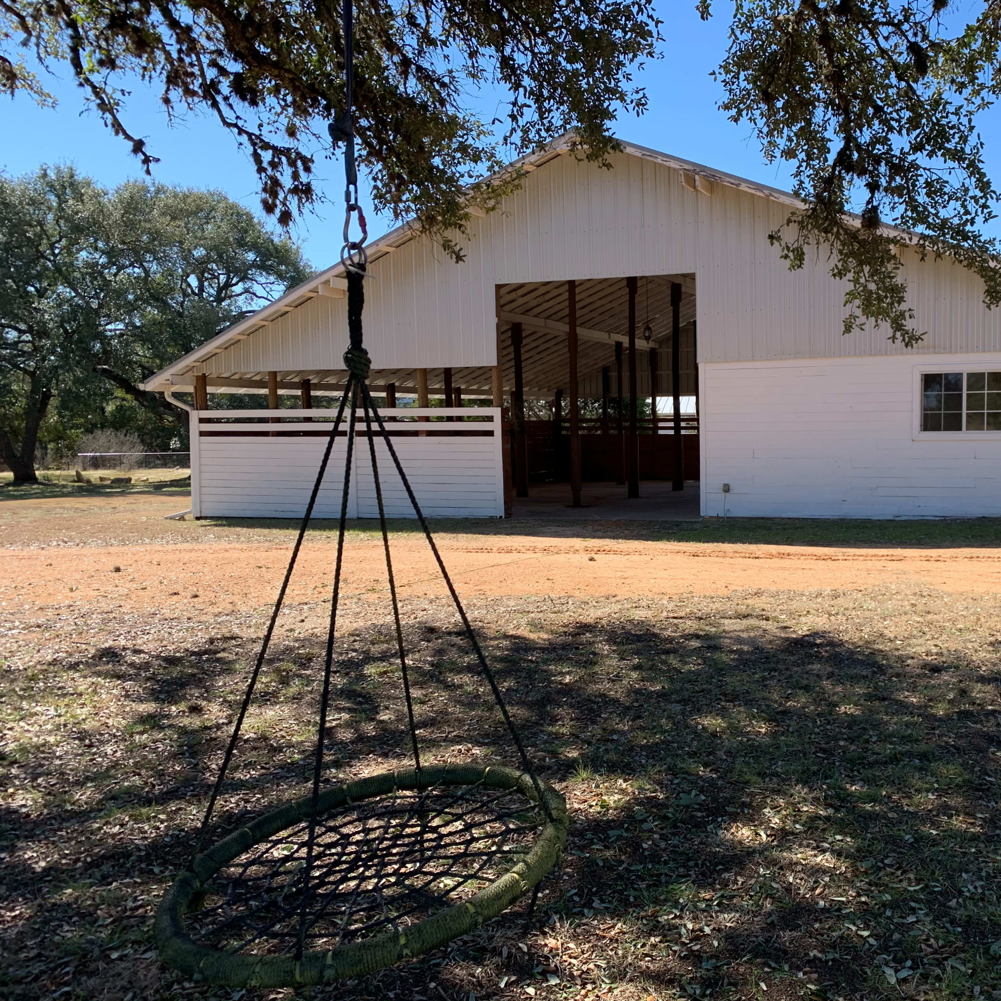 A white barn with large open doors stands beside a dirt path, and a hanging swing made of ropes and a circular seat is in the foreground.