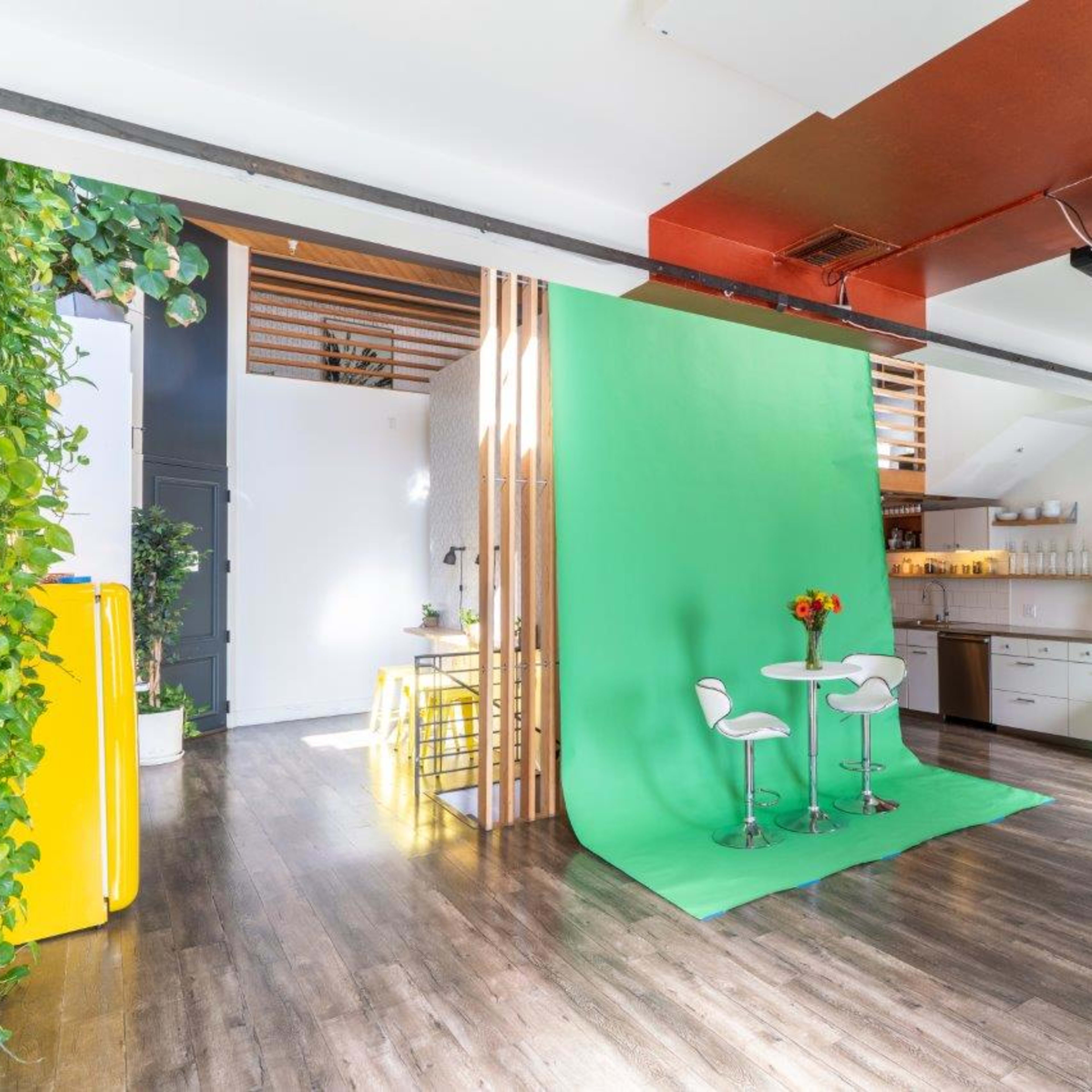 A bright, modern studio space with a green backdrop, two white stools, and a kitchen area, surrounded by plants and decorated with yellow pillows.