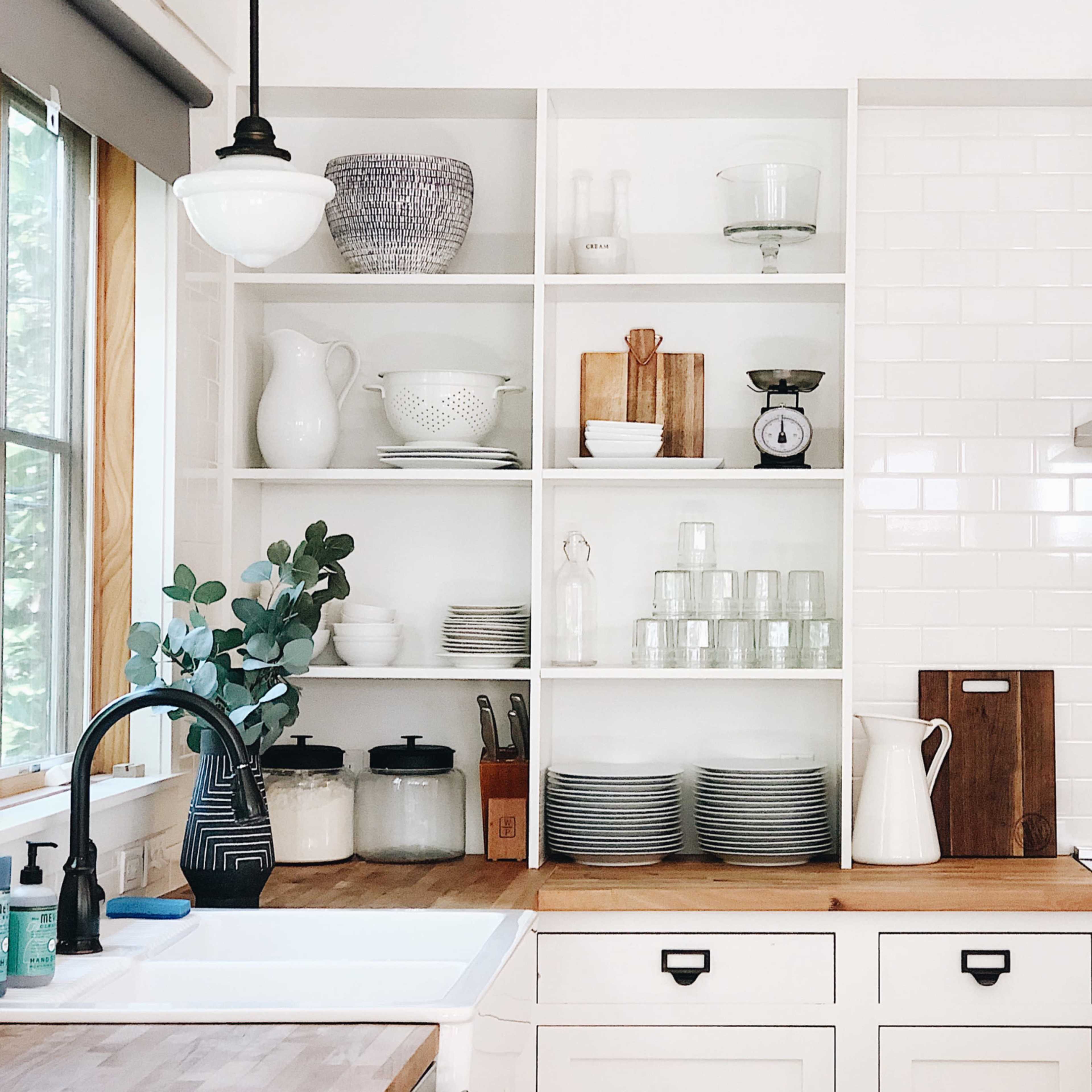 A modern kitchen with open shelving displaying various dishware, glassware, and kitchen tools above a wooden countertop and a stainless steel stove.