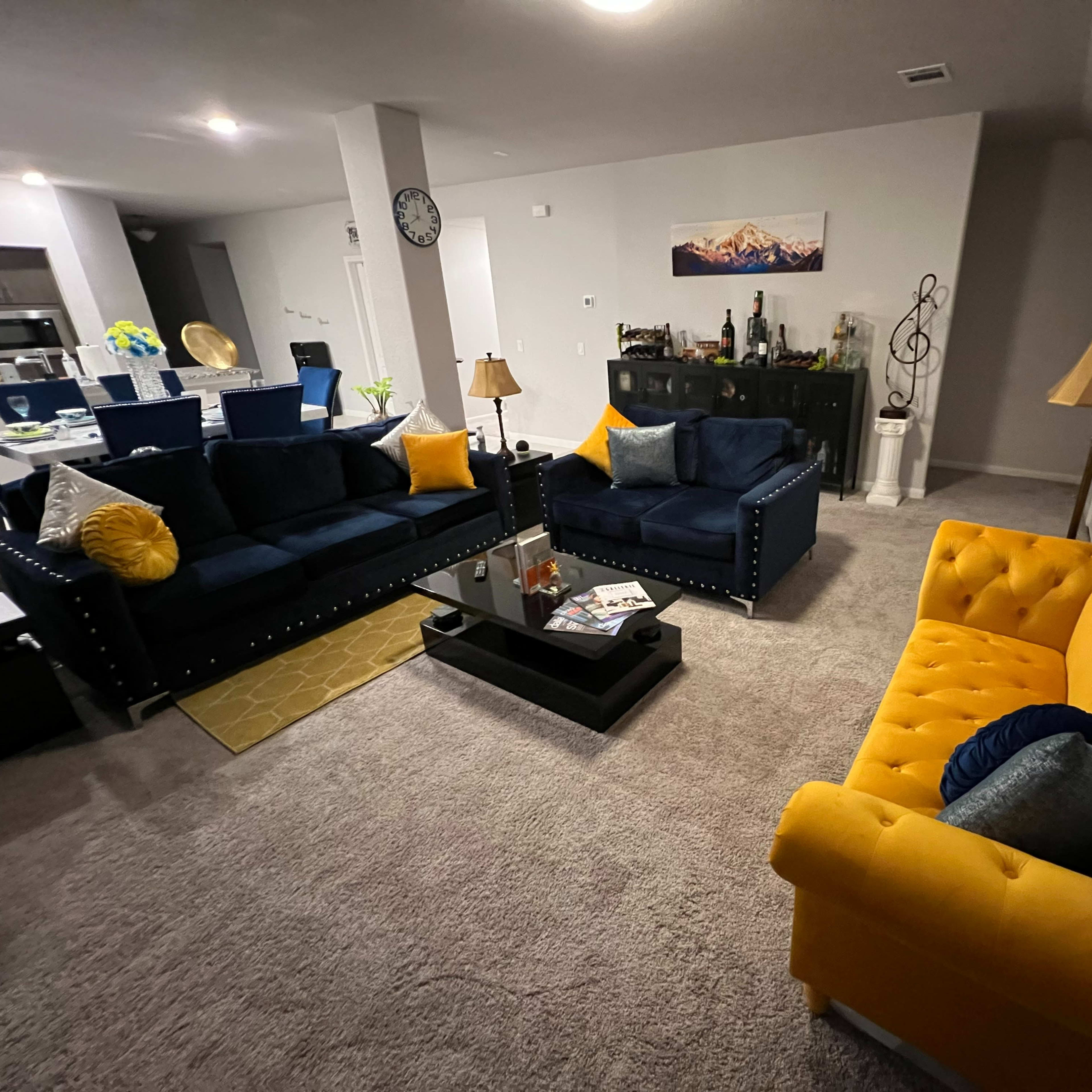 A living room with two navy blue sofas and a yellow loveseat arranged around a coffee table, with a dining area visible in the background.