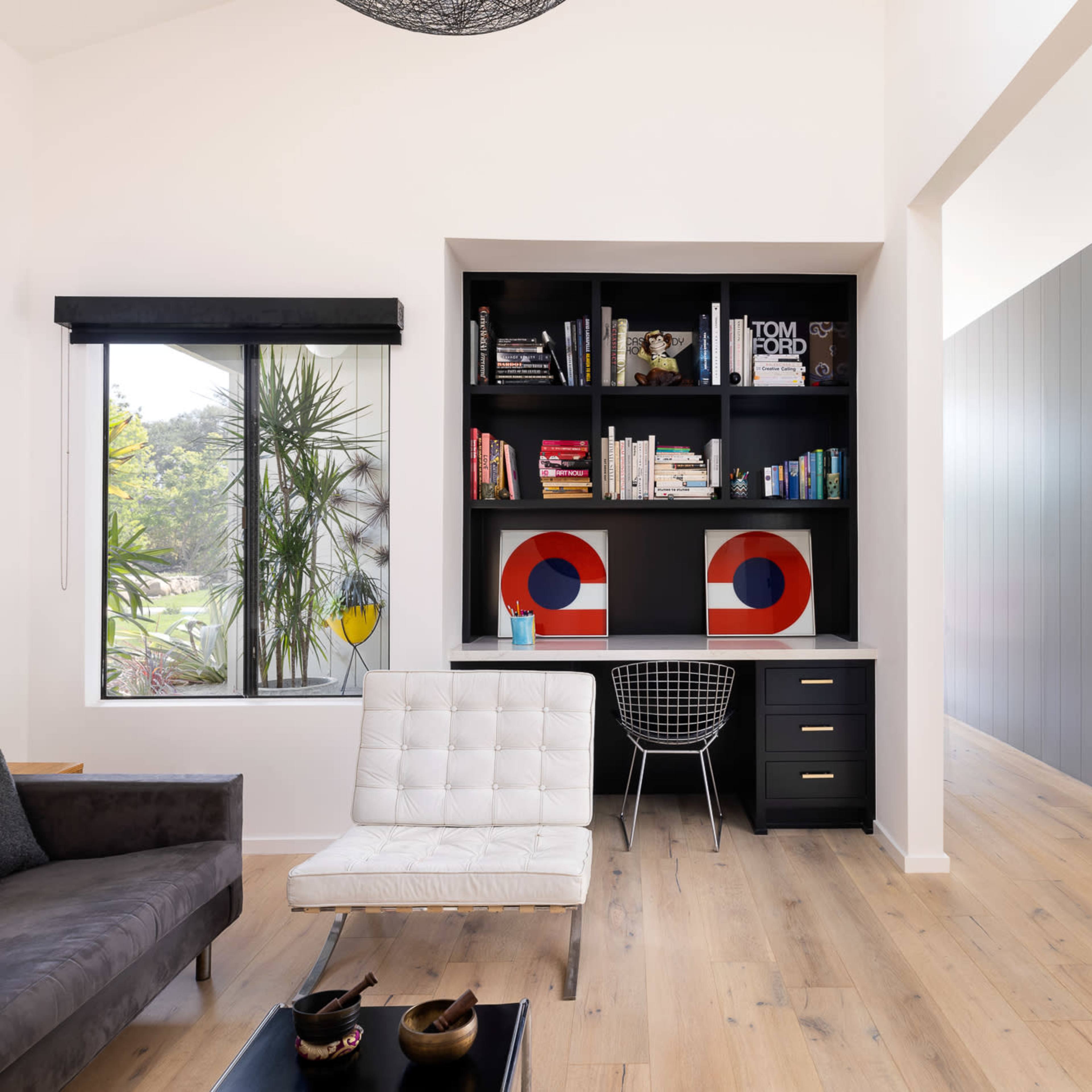A modern living space featuring a black bookshelf filled with books and decorative items, a white chair with a black wire base, and a cozy sofa positioned in front of a large window.