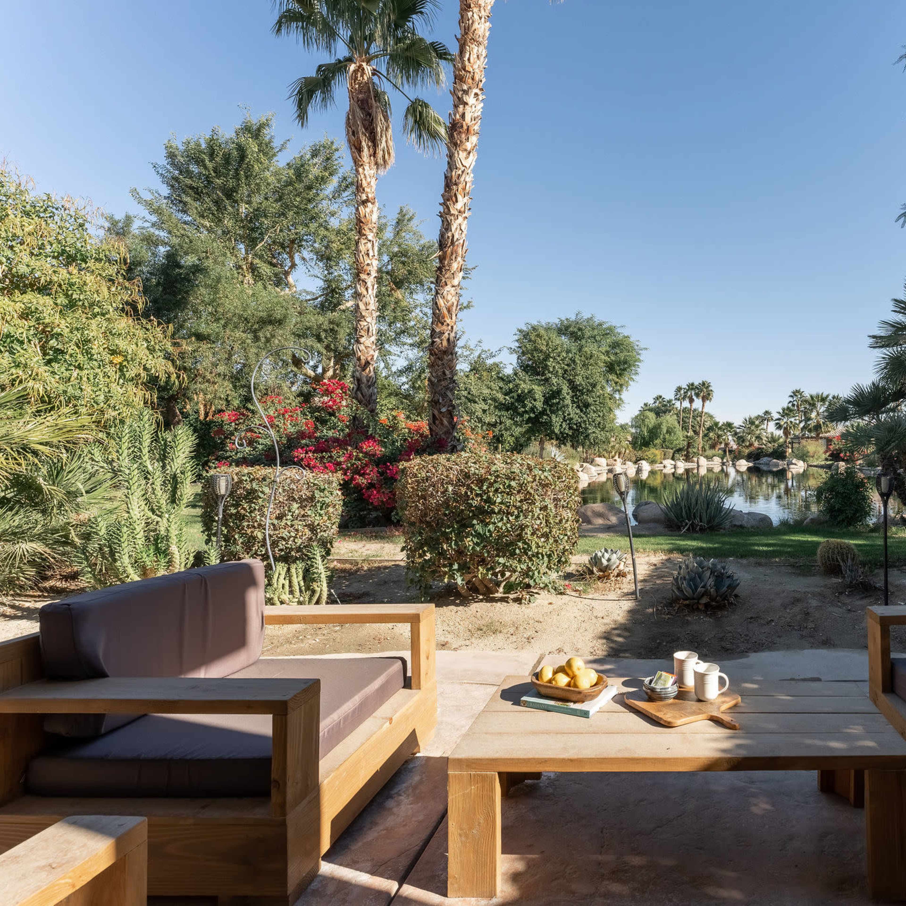 A backyard with two wooden chairs and a table, surrounded by various plants and palm trees, overlooking a tranquil water view.