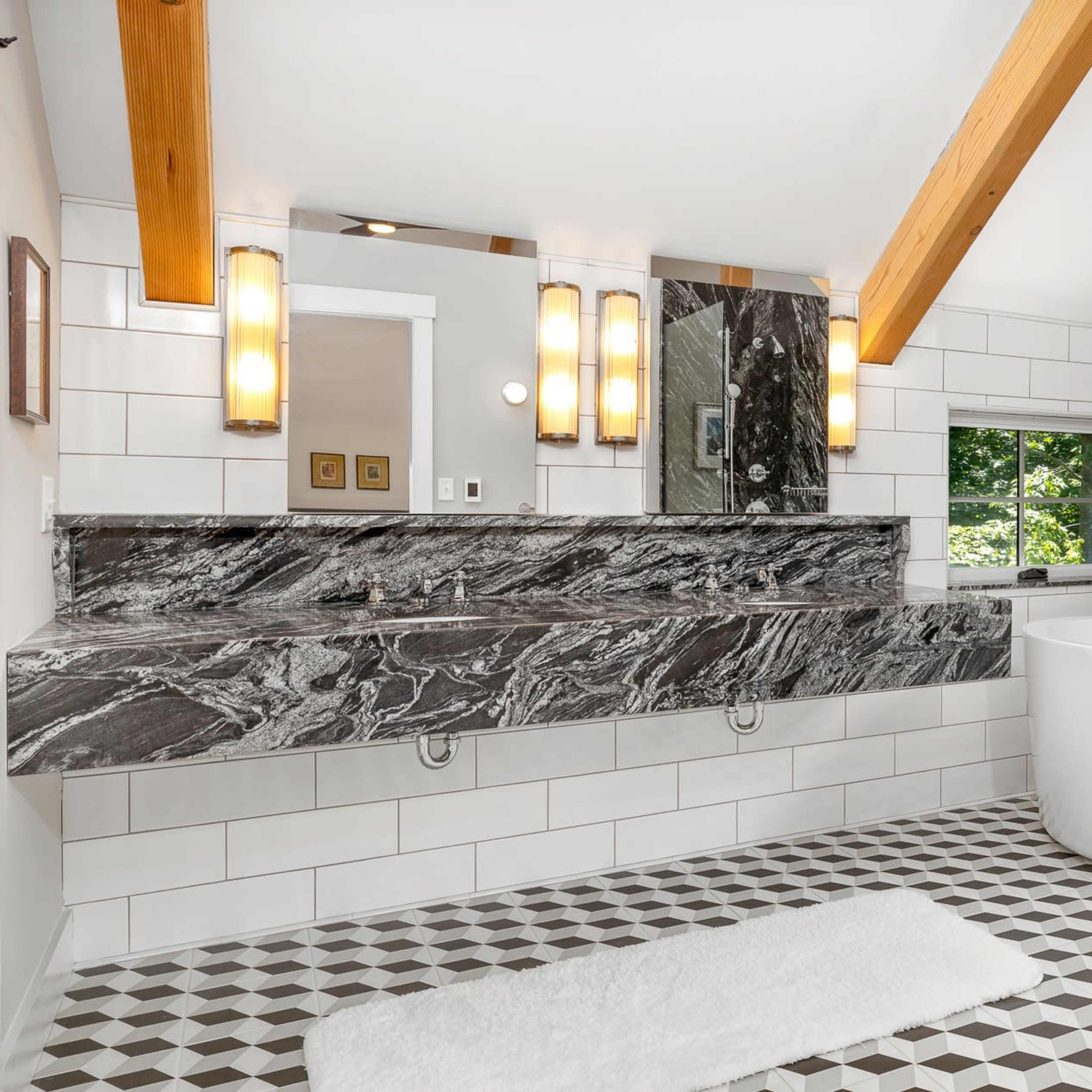 A modern bathroom featuring a gray marble countertop with two sinks, a round freestanding bathtub, and geometric patterned floor tiles.