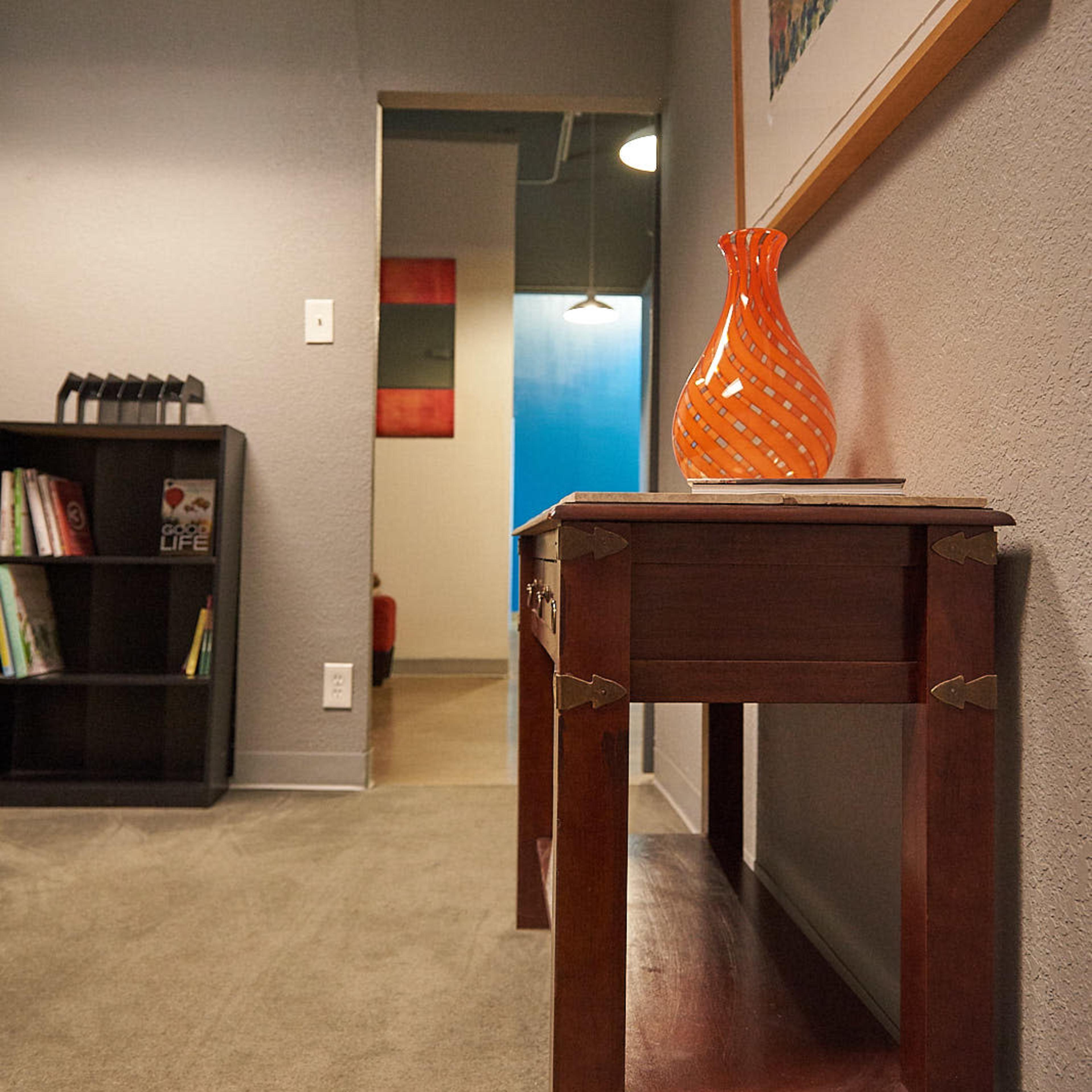 A wooden table with an orange vase on top is positioned in a hallway leading to rooms with colorful artwork.