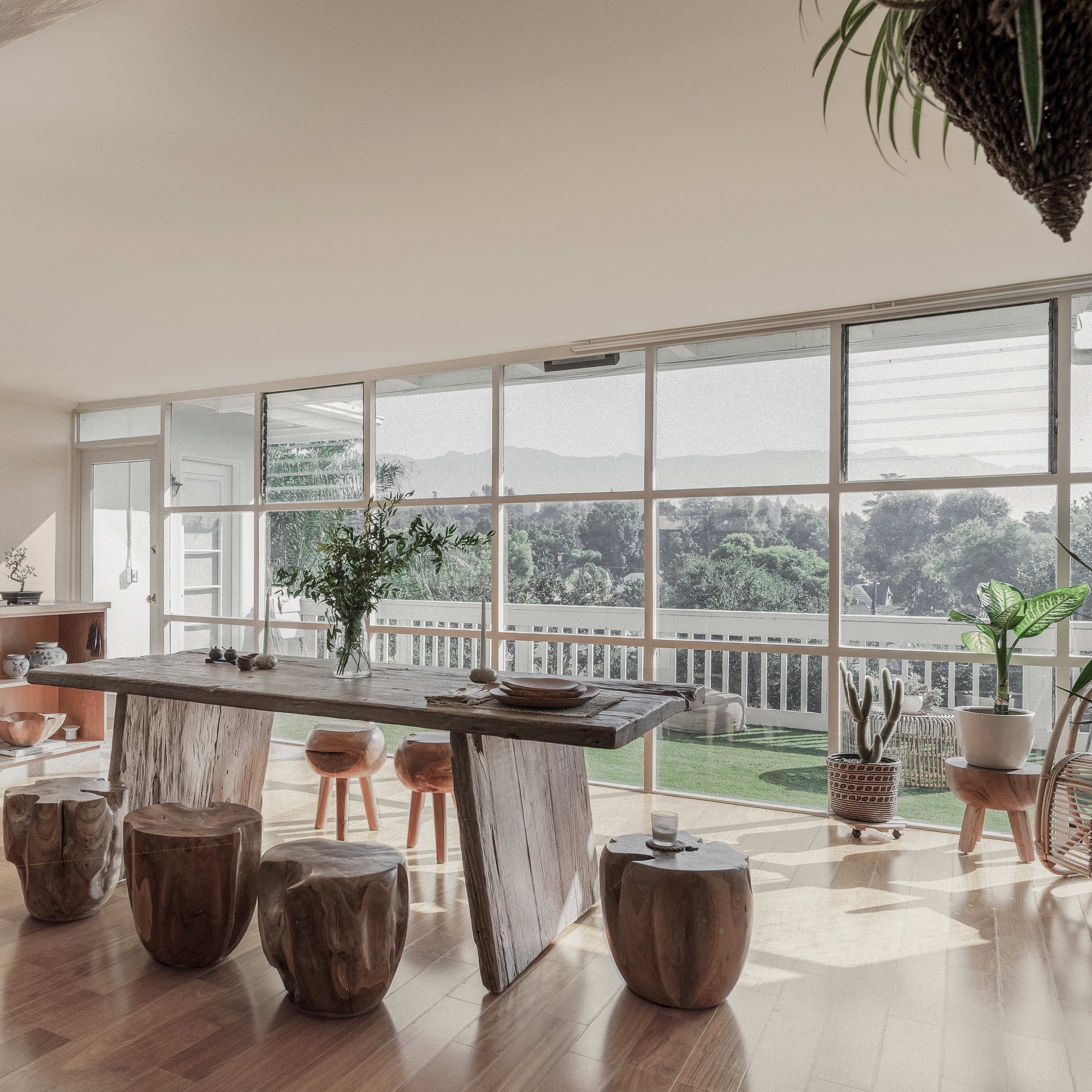 A spacious, well-lit room features a wooden dining table surrounded by tree stump stools, with shelves displaying various ceramic items and a view of greenery through large glass windows.