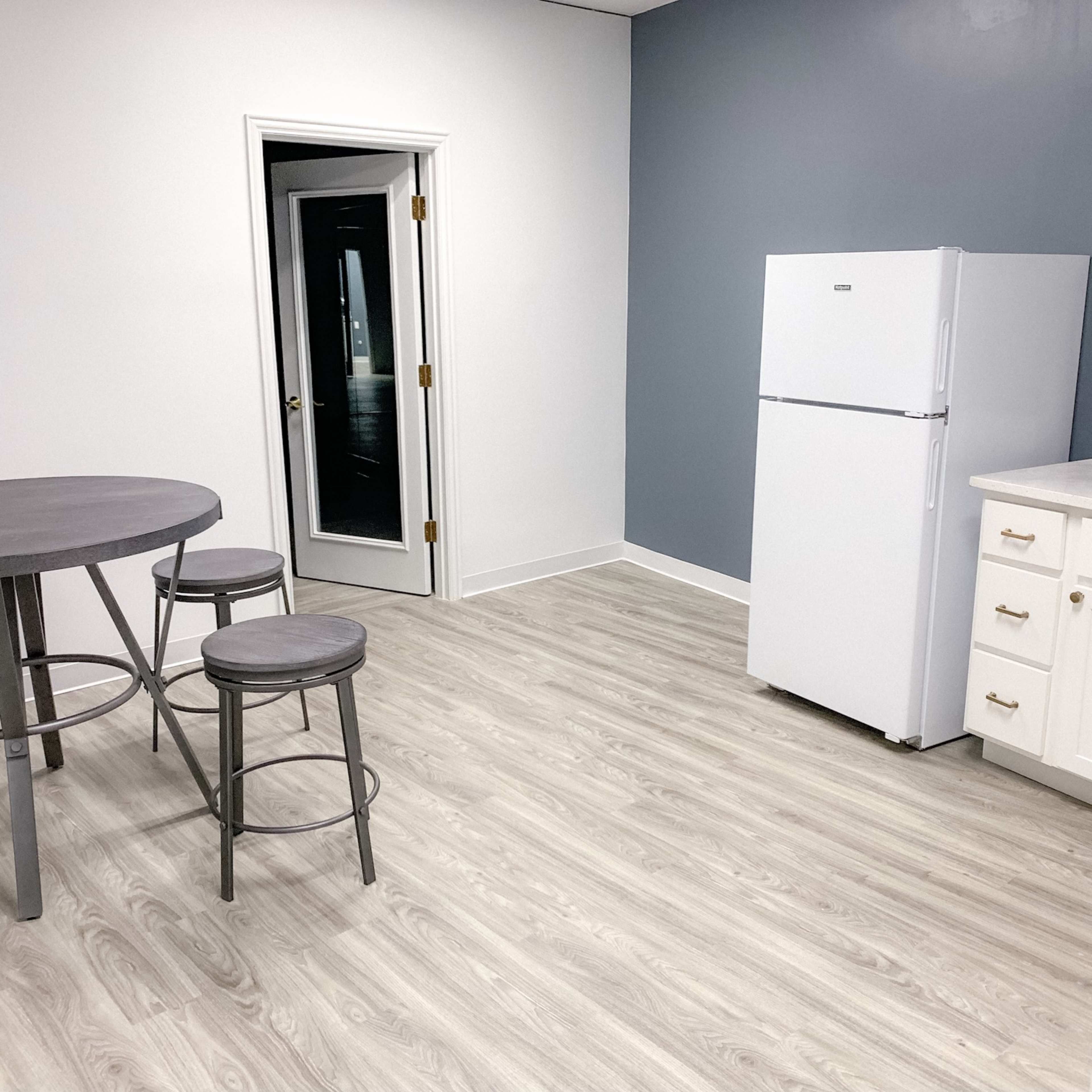 A simple kitchen area featuring a white refrigerator, a round dining table with stools, and a gray wall.