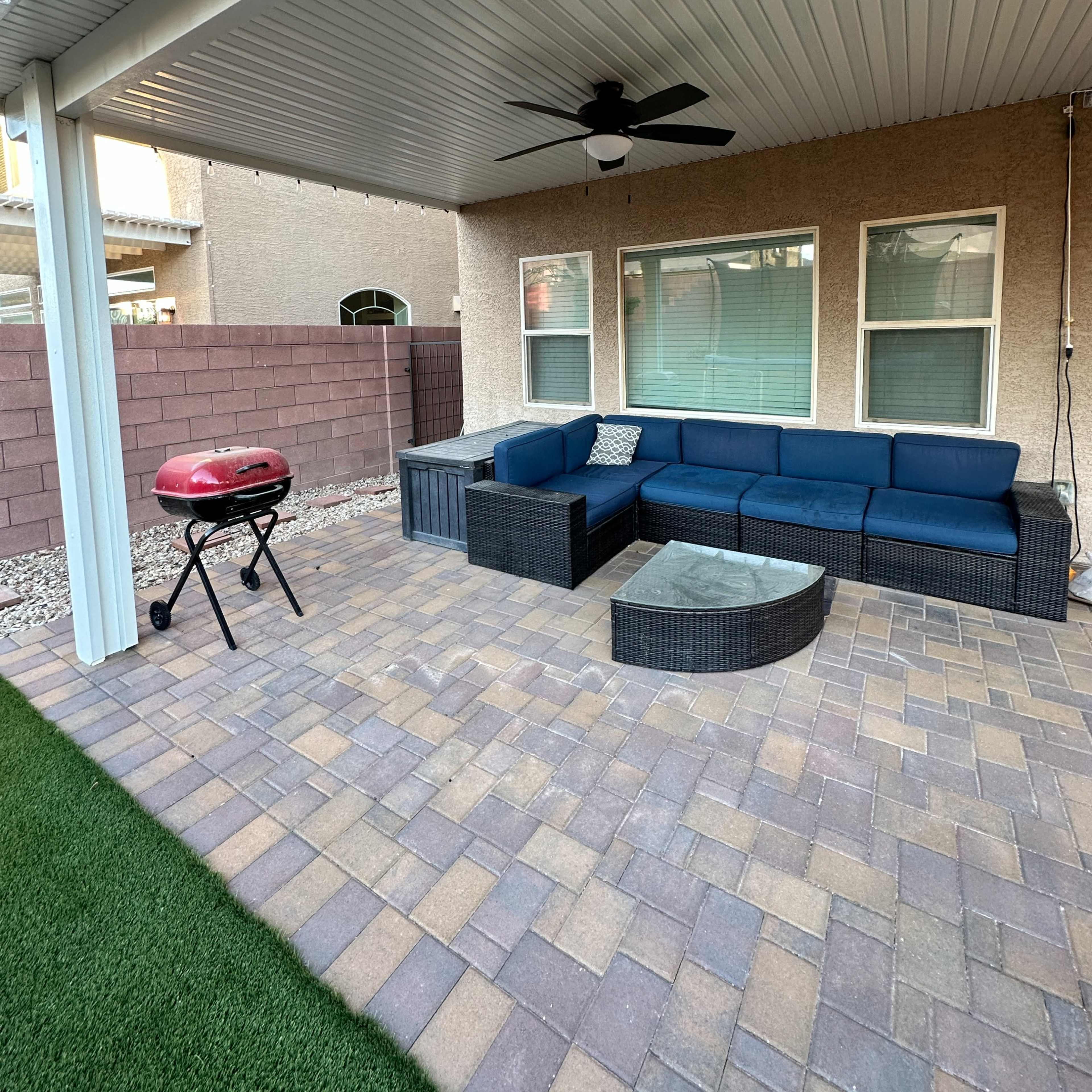 A covered outdoor patio with a blue sectional sofa, a glass coffee table, a grill, and pavers on the ground.