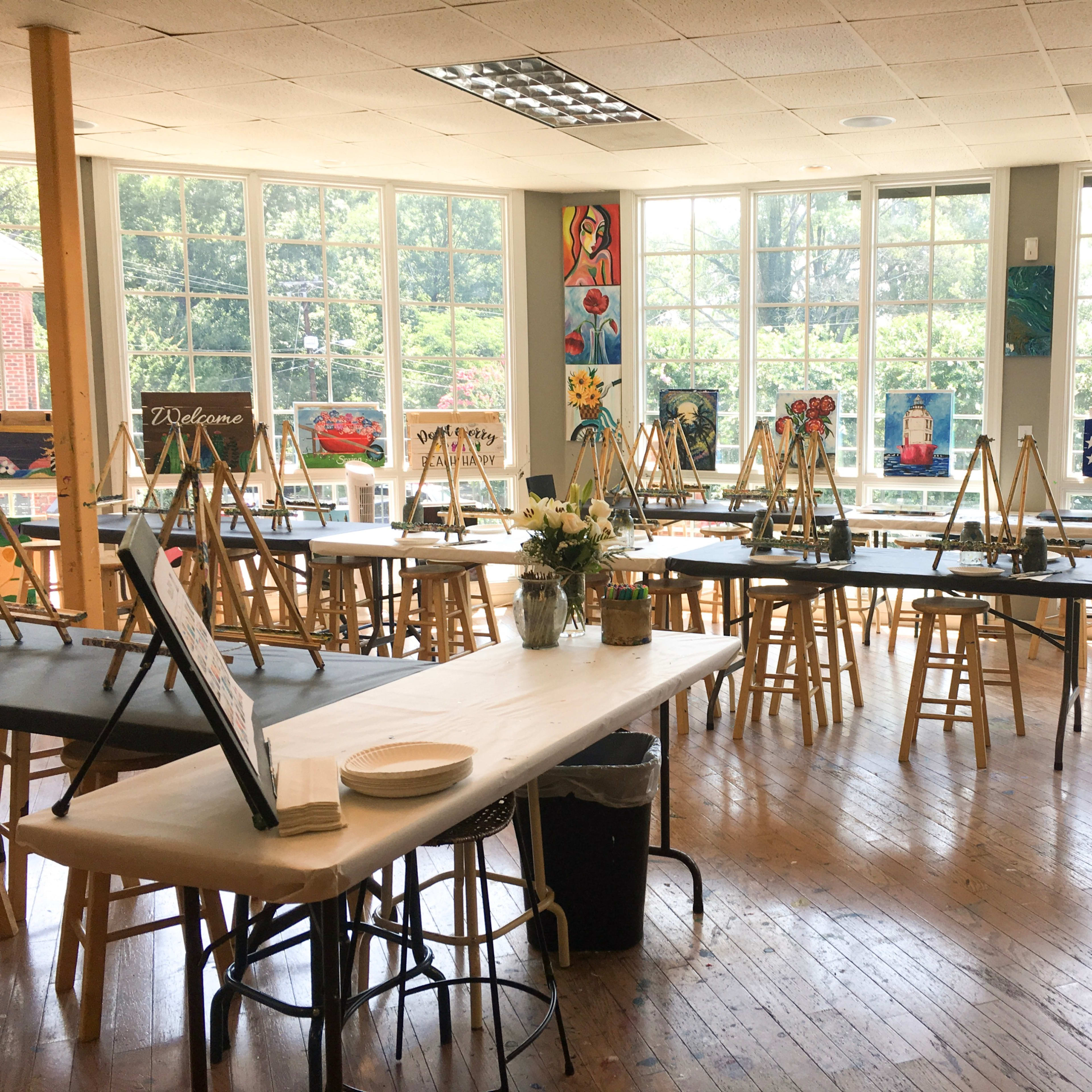 An art studio with numerous easels set up around tables, large windows letting in natural light, and artwork displayed on the walls.