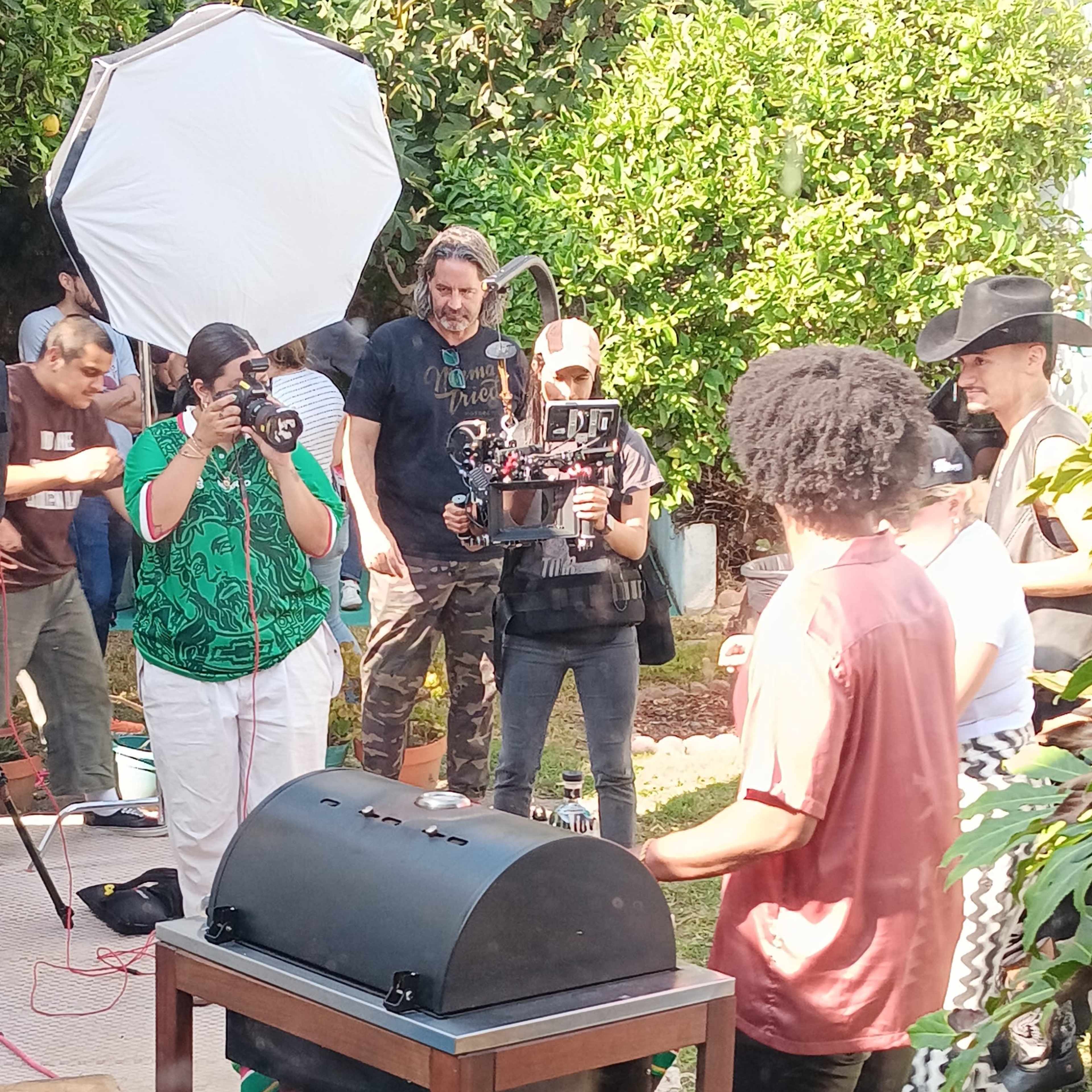 A group of people gathers around a grill in a backyard while several individuals film and take photos.