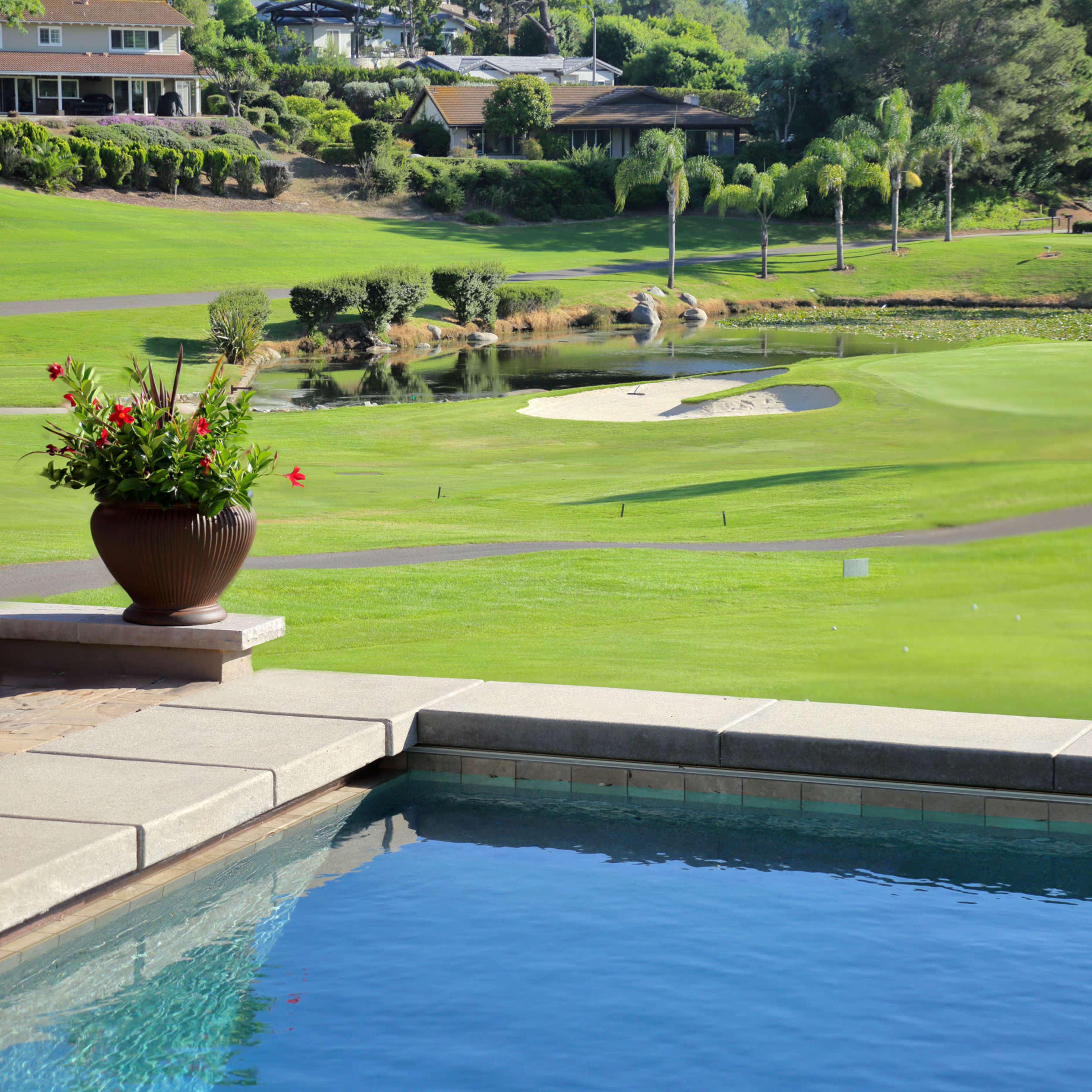 A swimming pool with a flower pot in the foreground overlooks a landscaped golf course with a pond and palm trees in the background.