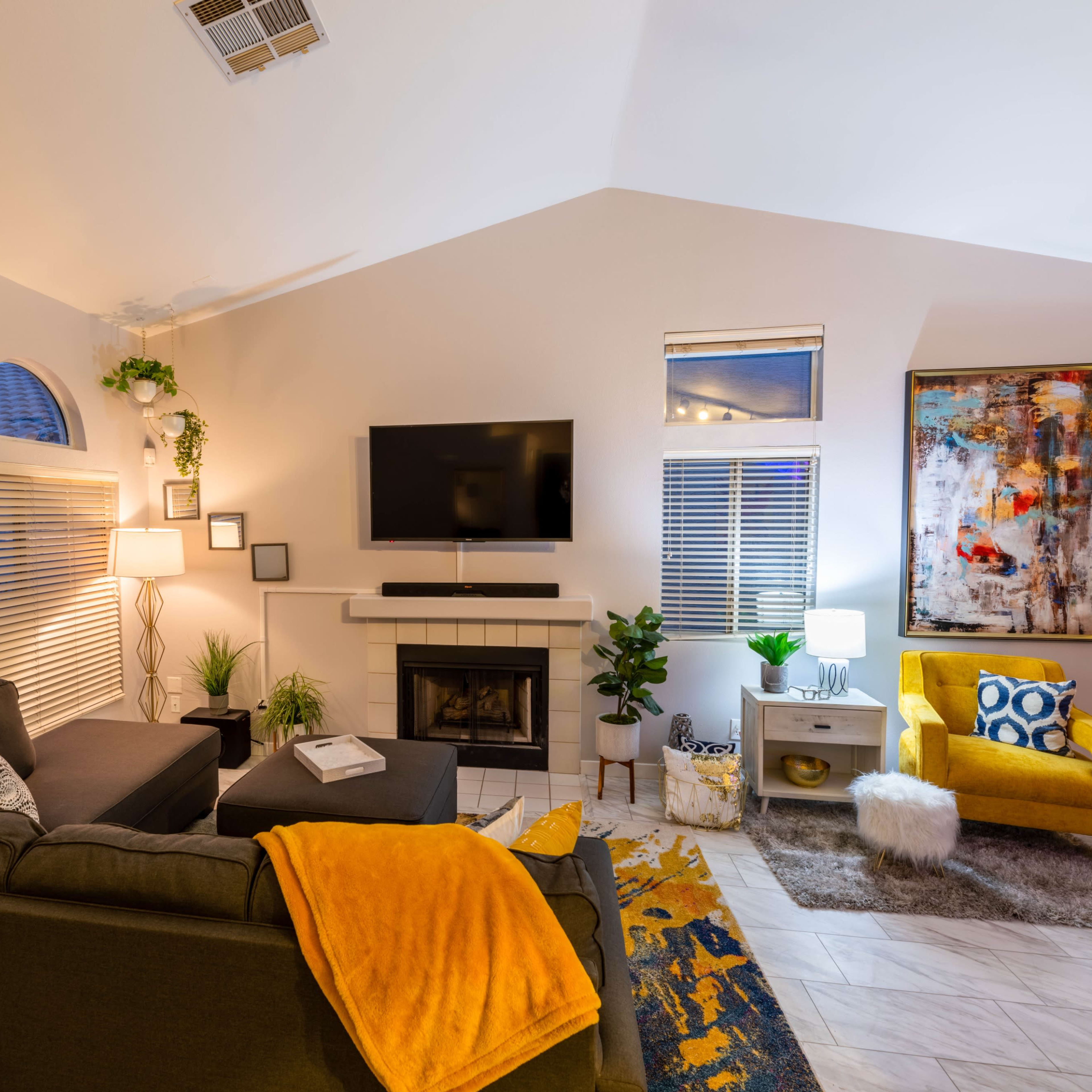 A modern living room features a gray sofa, yellow accent chair, fireplace, and a large wall-mounted television.