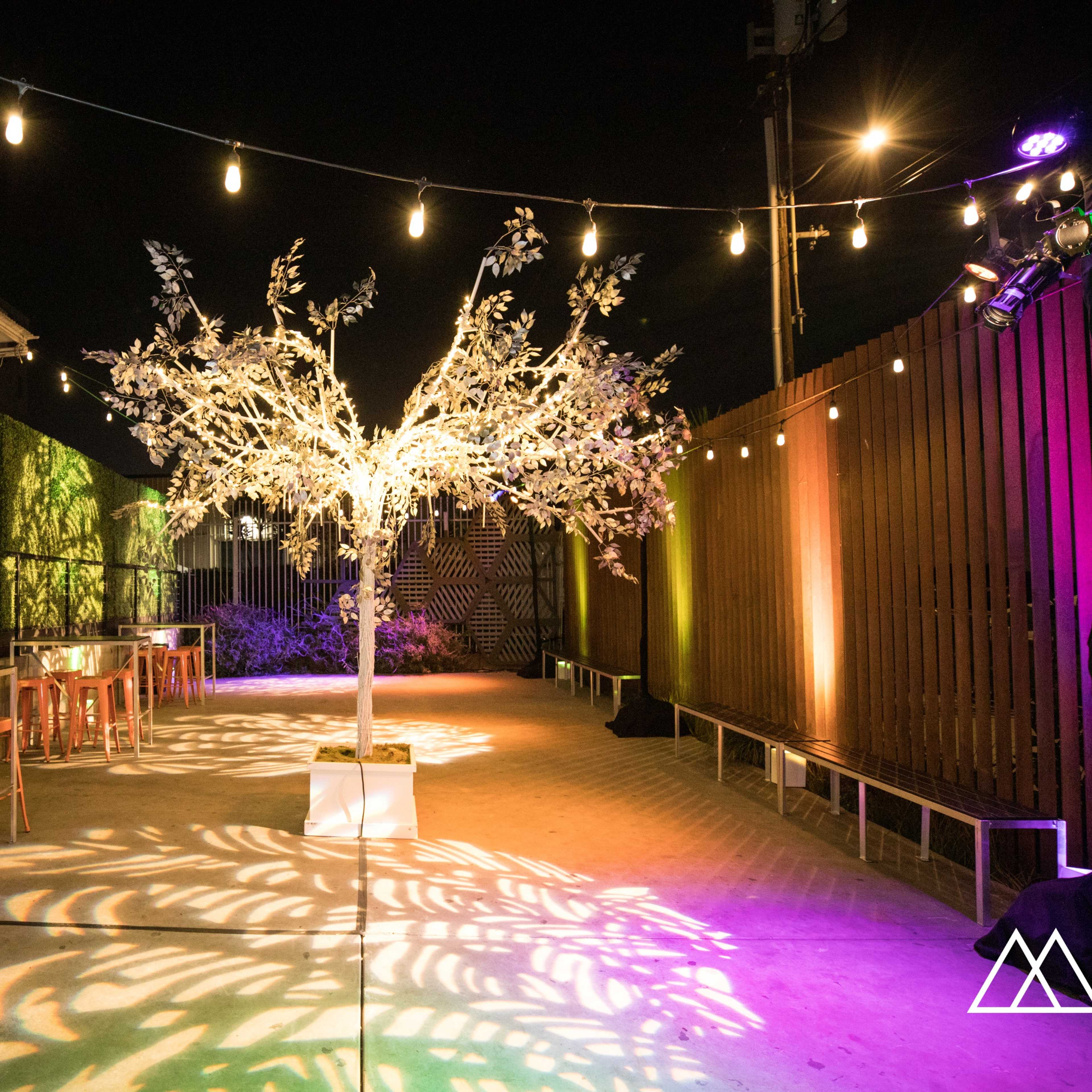 A lit outdoor event space featuring a decorative tree and several tables with chairs, surrounded by colorful lighting and greenery.