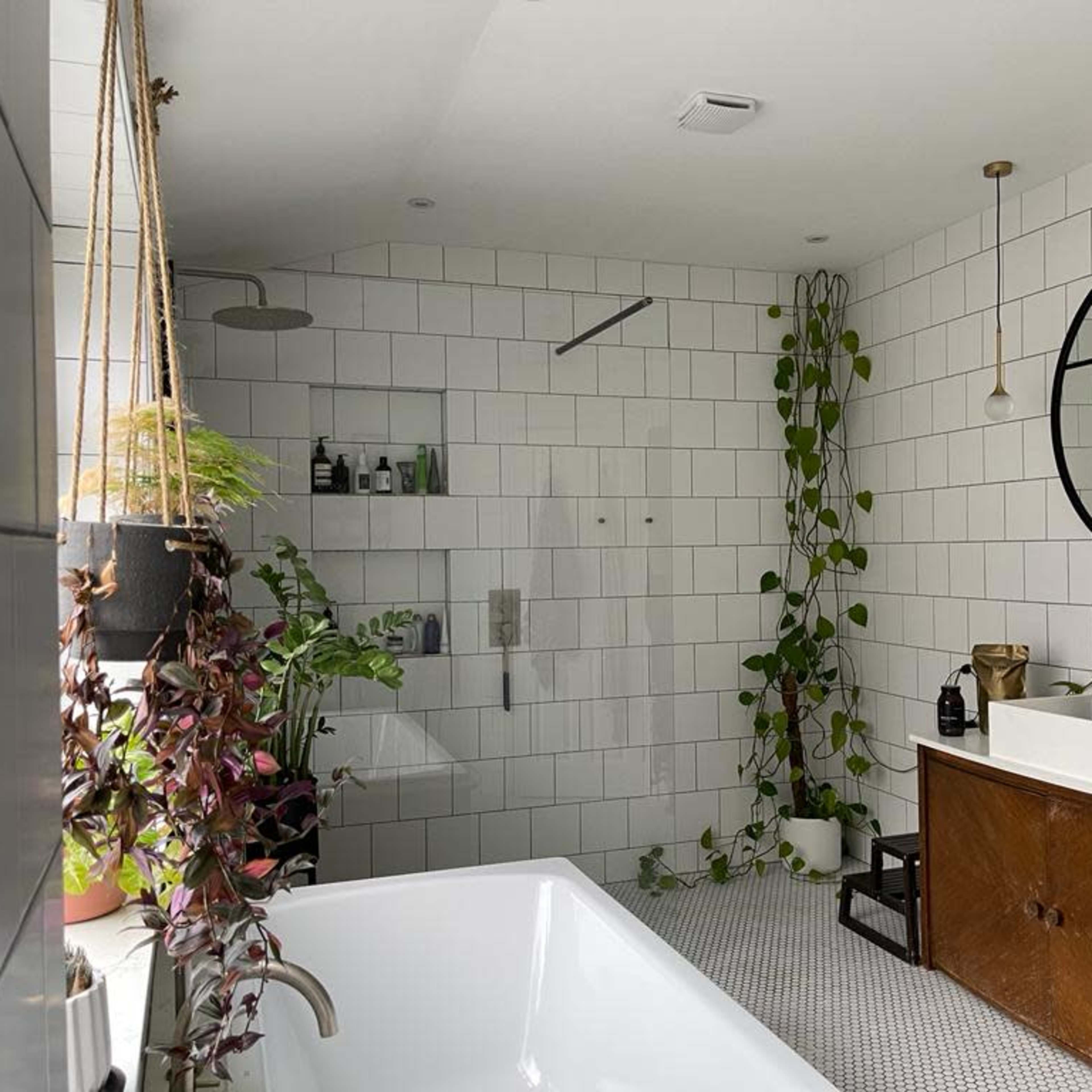 A modern bathroom features a freestanding tub, a large round mirror, plants, and a wooden vanity against a tiled wall.