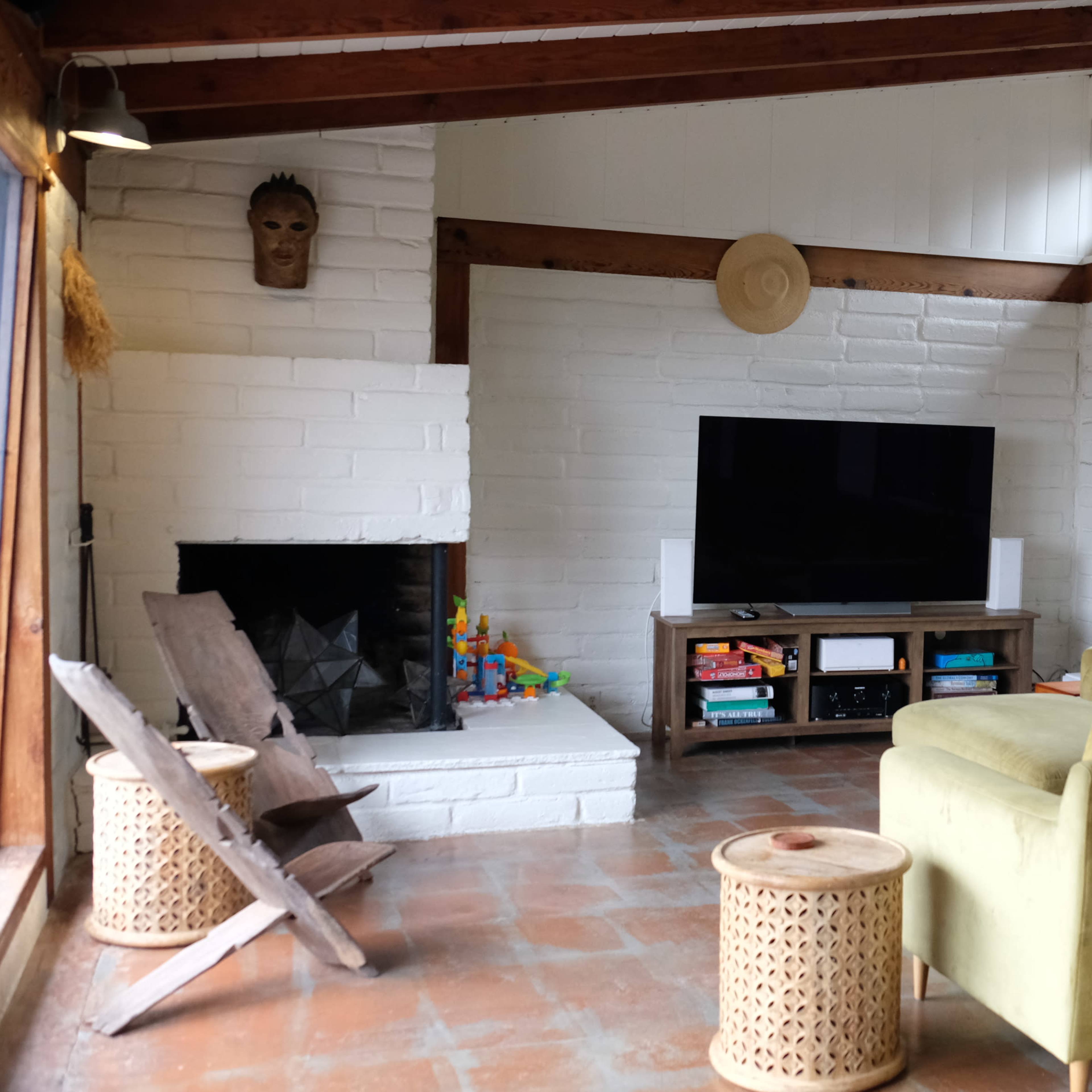 A living room with a green couch, a television on a wooden stand, and a fireplace, all set within a space featuring wooden beams and large windows.