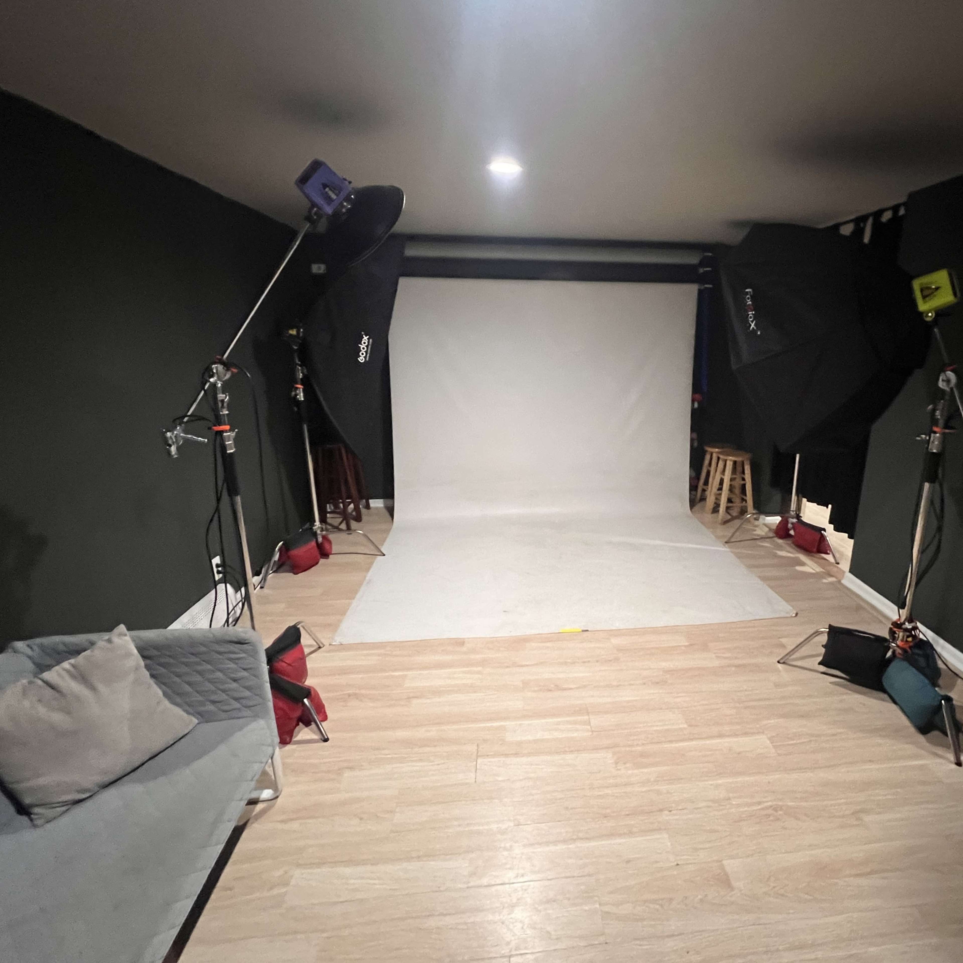 A photography studio with two large softbox lights, a white backdrop, and a gray sofa positioned on a wooden floor.
