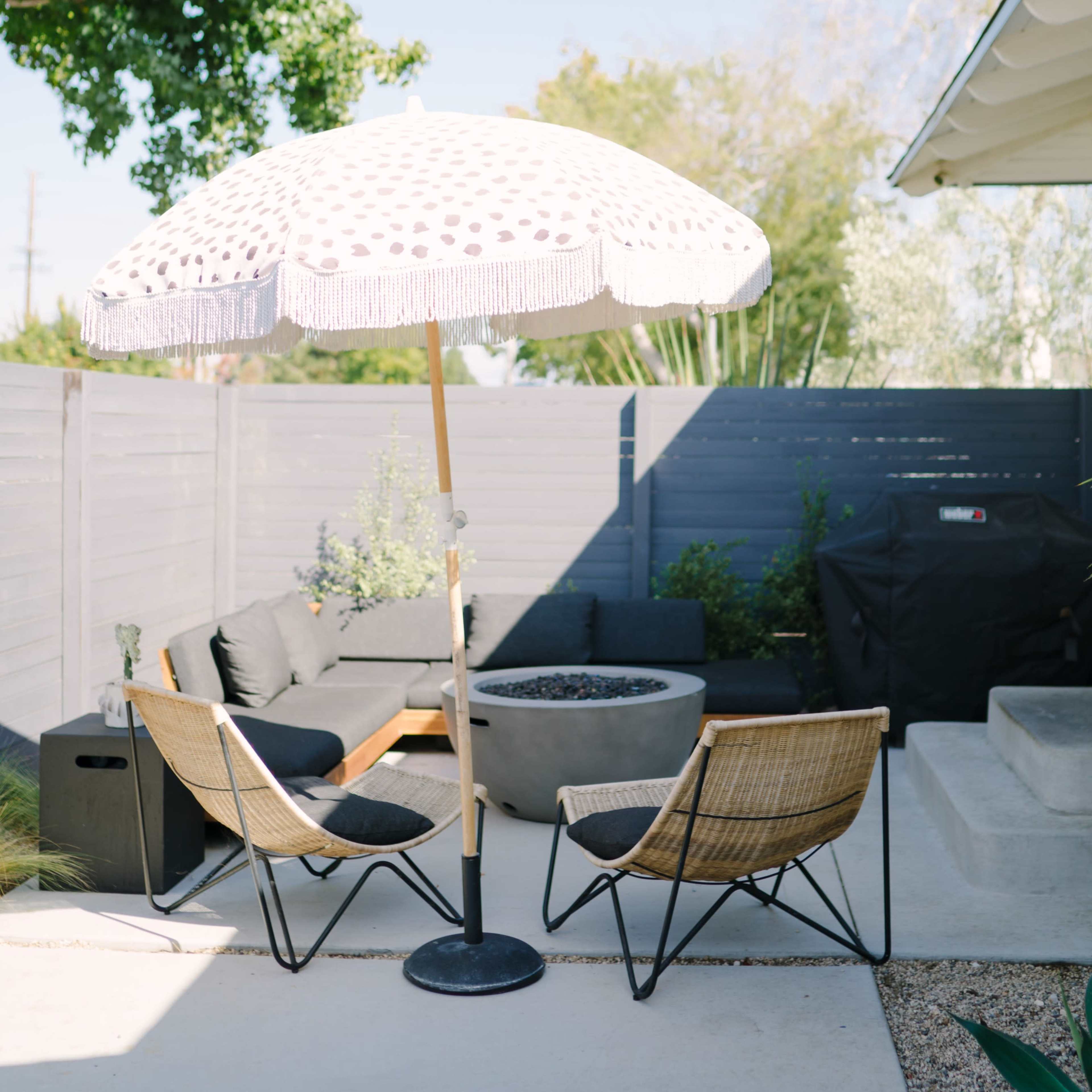 A patio area features two chairs under a large white umbrella, with a small table and a fire pit surrounded by greenery and a charcoal grill.