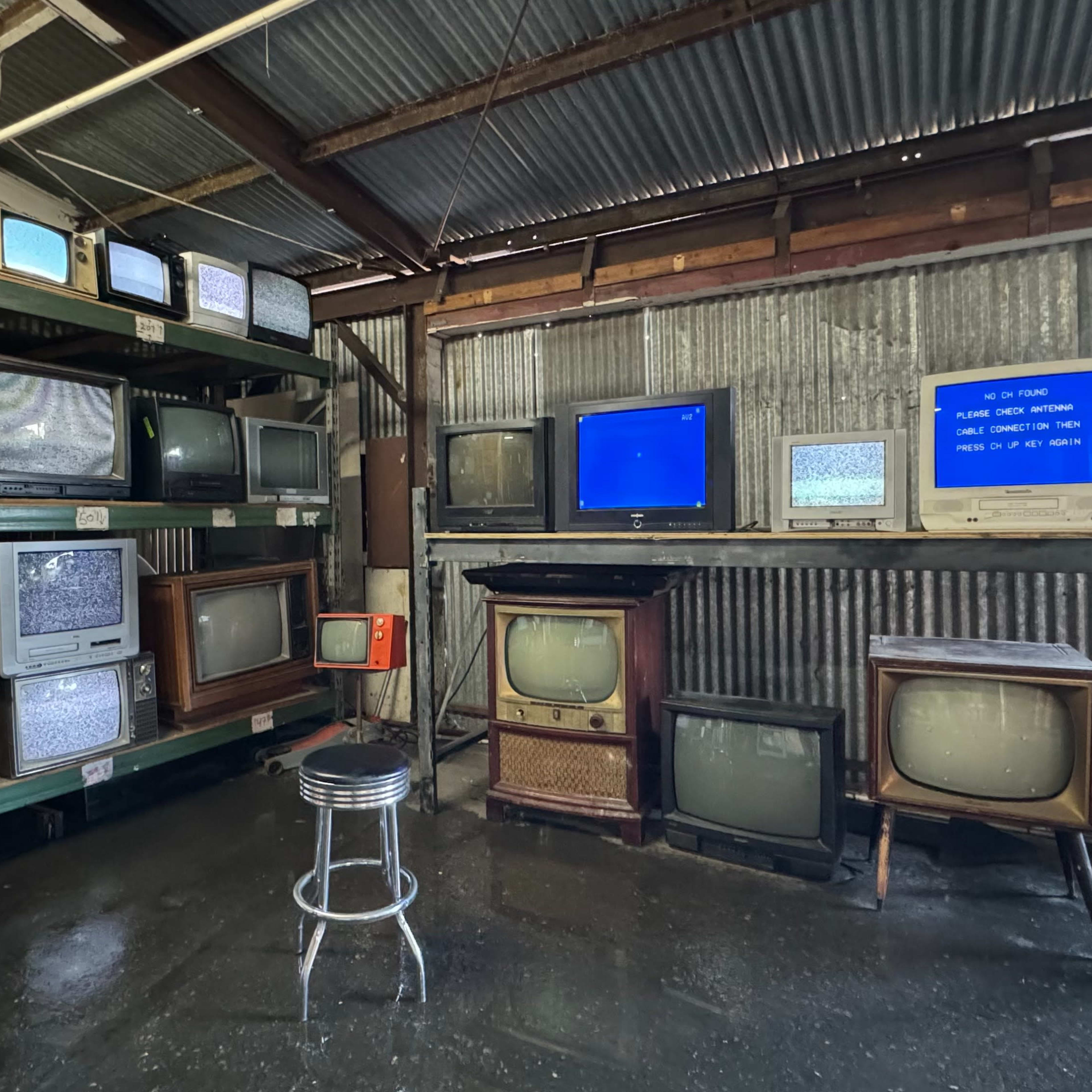 A warehouse setting with multiple vintage televisions arranged on shelves and a stool in front of them.