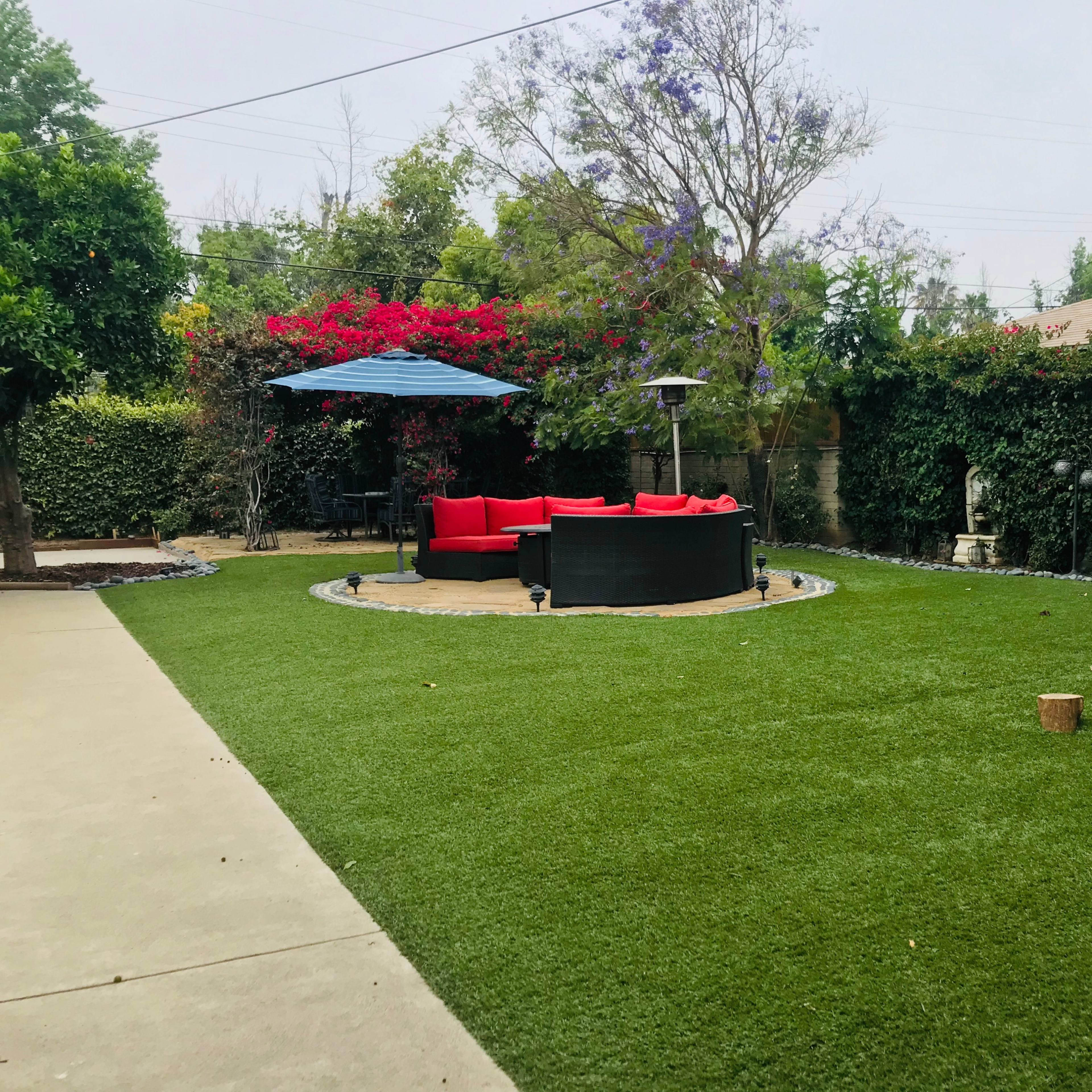 A landscaped backyard features a circular area of artificial turf with a black seating arrangement and red cushions, surrounded by greenery and flowering plants.